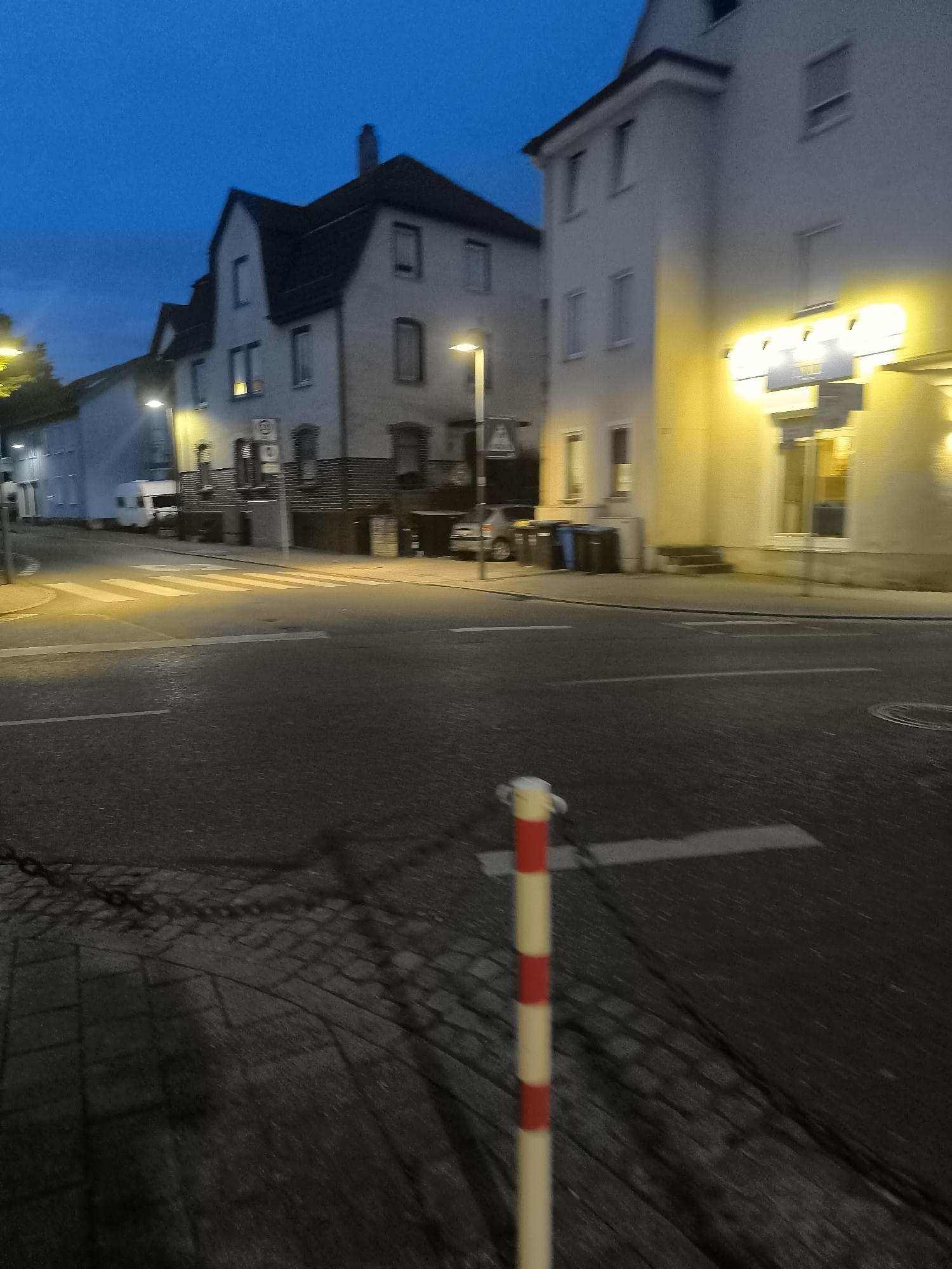 The image captures a street scene at dusk or night in an urban environment. In the foreground, a red and white striped bollard stands on the right side of the frame. The street itself is paved, with visible lane markings and a pedestrian crossing marked by white stripes. To the left of the street, a cobblestone pavement is visible.
In the background, two buildings are prominent. The building on the right is a multi-story structure with a bright yellow illuminated sign above its entrance. The building on the left appears to be a residential or commercial building with multiple windows and a sloped roof. A streetlamp illuminates a portion of the street and the building on the left. A white camper van is parked in front of the building on the left, and several trash bins are visible near the building on the right. The sky is a deep blue, indicating low light conditions.
