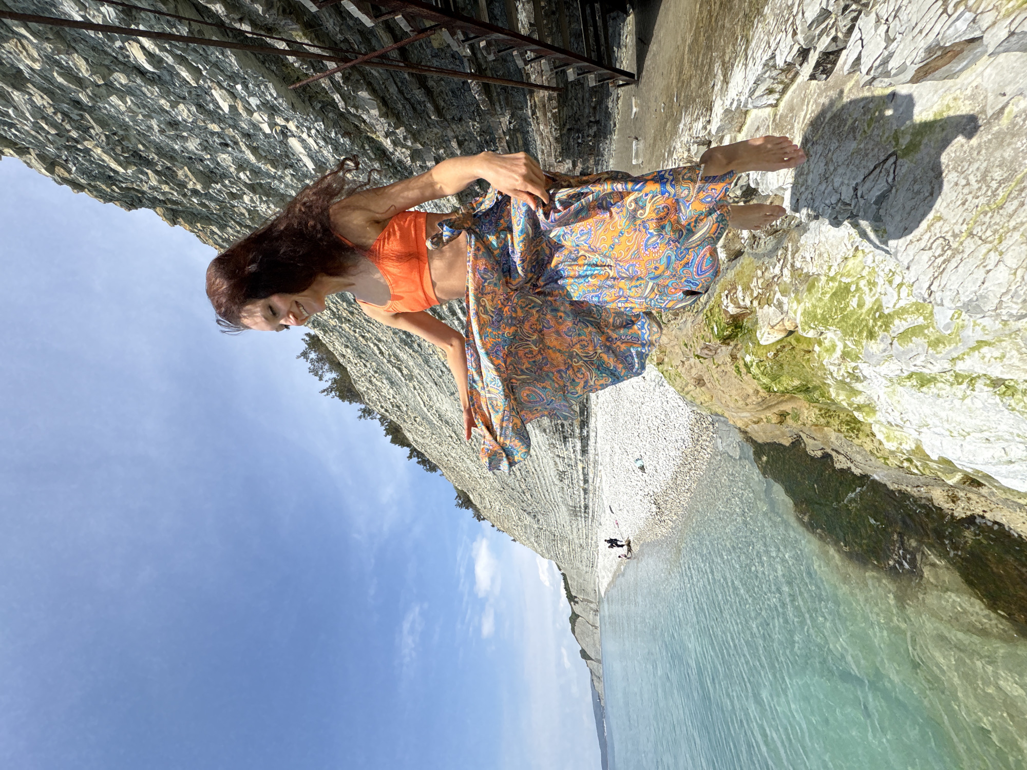 The image, when correctly oriented, depicts a woman posing on a sunny, rocky beach alongside a tall, layered cliff. The woman, with long, dark, wavy hair, smiles gently, looking slightly downward. She is wearing an orange cropped top and a flowing, multi-colored skirt with an intricate blue and orange paisley pattern. Her bare feet are visible on the light-colored, pebbly shore.
Behind the woman and extending across the left side of the frame is a striking cliff face, composed of distinctive horizontal layers of light gray and white sedimentary rock. Some green algae or moss clings to the rocks nearer the water's edge. At the top of the cliff, a metallic structure, possibly part of a staircase or railing, is visible.
To the woman's left, the clear, turquoise water of the sea stretches into the distance. The coastline continues into the background, revealing more white cliffs and a wide, pebbly beach where a few small figures of people are visible, along with what appears to be a small inflatable boat or paddleboard in the water. The sky above is a bright, clear blue with scattered white clouds, indicating a sunny day. Strong sunlight casts distinct shadows on the rocky ground. The scene evokes a tranquil coastal setting. No visible text is present.