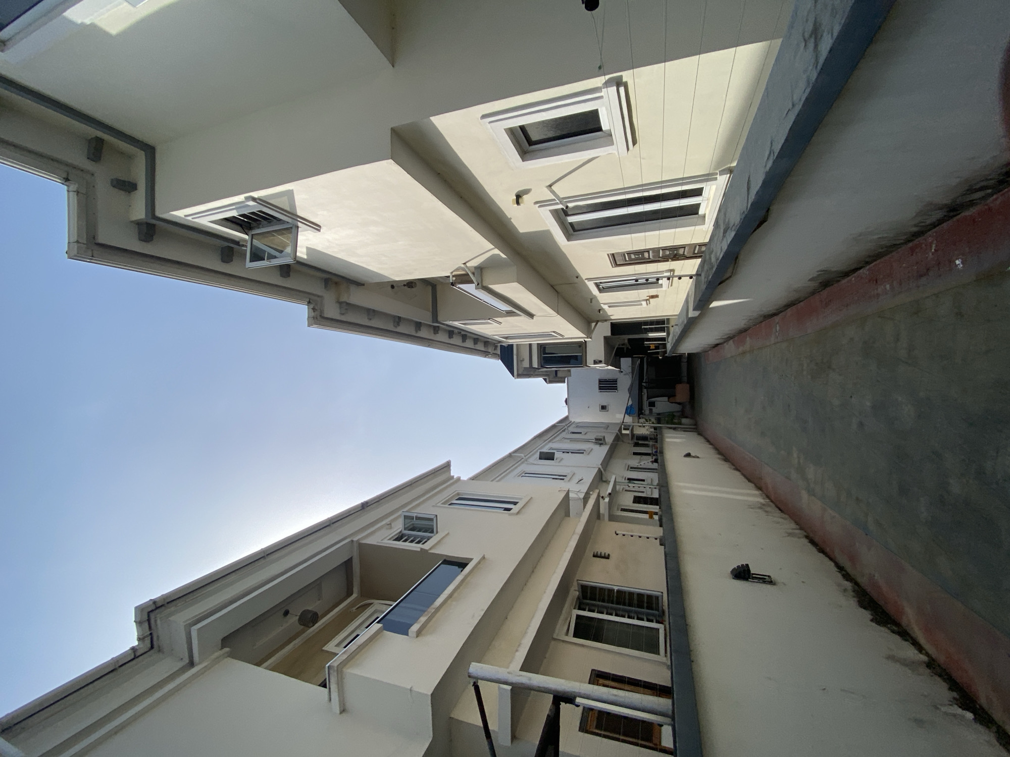 An upward-angled view shows two buildings facing each other in Lagos, Nigeria. The buildings are painted off-white and have multiple windows of varying sizes. One building appears taller than the other. A narrow pathway runs between them, and the sky is visible above. The image seems to be taken in a narrow alleyway. The weather looks sunny, and there is nothing in the foreground. The general environment is urban. No people are visible.