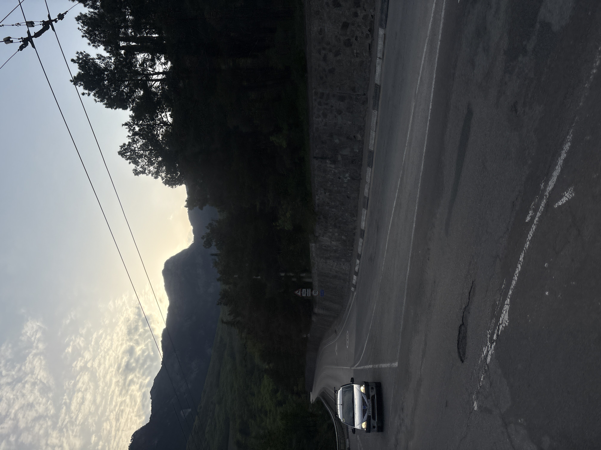 The image shows a mountain road near Массандровский поселковый совет, Ukraine. A white van with its headlights on is driving on the right lane of the road. The road curves slightly to the left, with a stone wall on the left side. Beyond the wall, a steep, tree-covered mountainside rises. The sky is partly cloudy, with the sun setting behind the mountains in the distance. Overhead power lines are visible in the top left corner of the image. The road surface appears worn, with some cracks and patched areas.