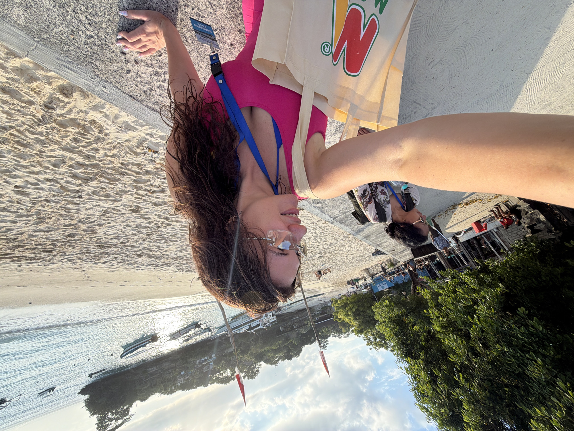 A woman in a pink top and blue bikini is taking a selfie on a sandy beach in Nusa Penida, Indonesia. She is wearing clear-rimmed glasses and a blue lanyard with a pass attached. A canvas tote bag with a red and yellow logo is slung over her shoulder. To her left, another woman in a floral shirt is partially visible. The background shows a beach with scattered boats on the turquoise water, lush green trees, and buildings in the distance. Two red flags on poles stand on the beach. The sun is setting, casting a warm glow on the scene.