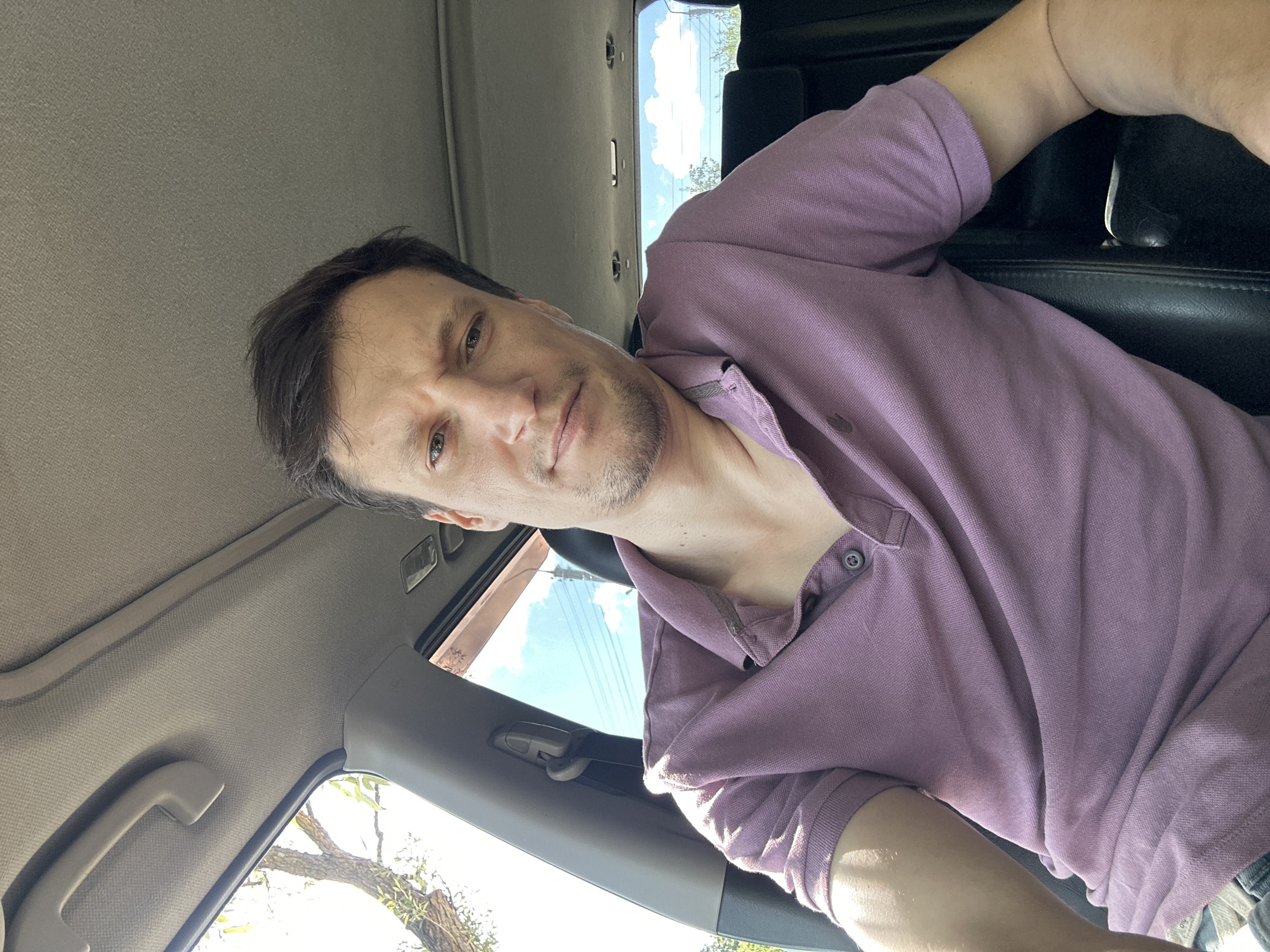 A man is sitting in the passenger seat of a car, looking at the camera. He is wearing a purple polo shirt and has short, dark hair. The car appears to be parked outdoors, with trees and a blue sky visible through the windows. The interior of the car is dimly lit, with the man's face illuminated by sunlight. The image was taken from a slightly low angle.