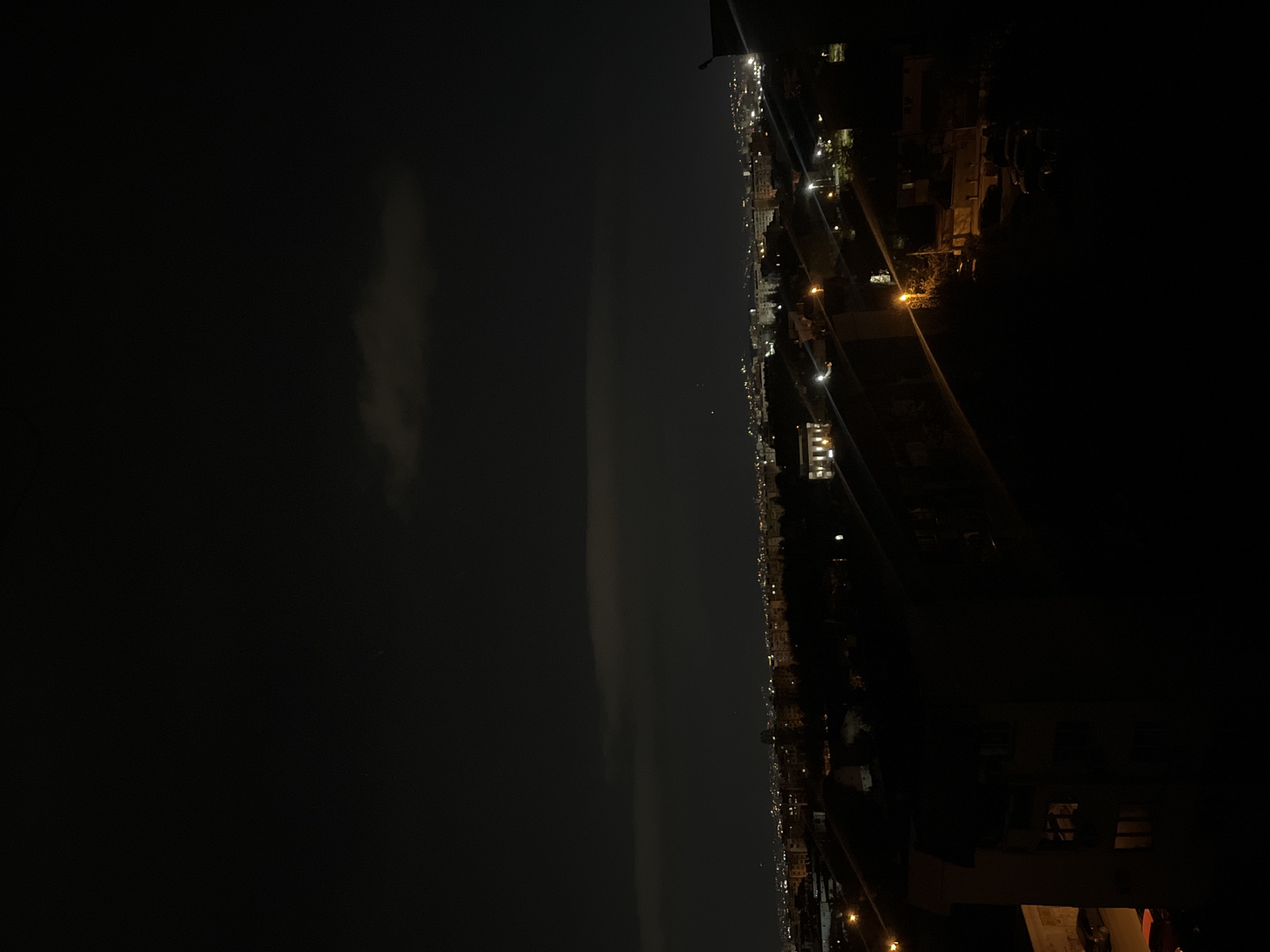 This media file presents a panoramic, elevated view of a city at night, specifically Sofia, Bulgaria, captured with a vertical orientation (rotated 90 degrees clockwise). When viewed correctly, the scene depicts a vast urban landscape illuminated by numerous artificial lights under a dark night sky.
The lower half of the image (right side in the provided vertical orientation) is dominated by the cityscape. A dense array of warm-toned lights from buildings, streetlights, and possibly vehicle traffic stretches horizontally across the frame, creating a striking pattern against the darkness. These lights delineate the contours of a sprawling urban area, indicating numerous structures and active thoroughfares. Some individual buildings are faintly visible, illuminated by their own internal and external lights.
The upper half of the image (left side in the provided vertical orientation) shows the night sky. It is predominantly dark, though some subtle, faint cloud formations or atmospheric haze are discernible, particularly a long, wispy band extending horizontally across the mid-sky area. There are no prominent stars or celestial bodies visible, suggesting either light pollution from the city below or cloud cover.
The perspective is high, likely from a tall building or an elevated vantage point overlooking the city, offering a wide and distant view of the urban expanse. No specific activities or events involving people are visible; it is a static landscape scene. There is no discernible text within the image. The overall impression is one of a vast, tranquil city illuminated by its own glow under the night sky.