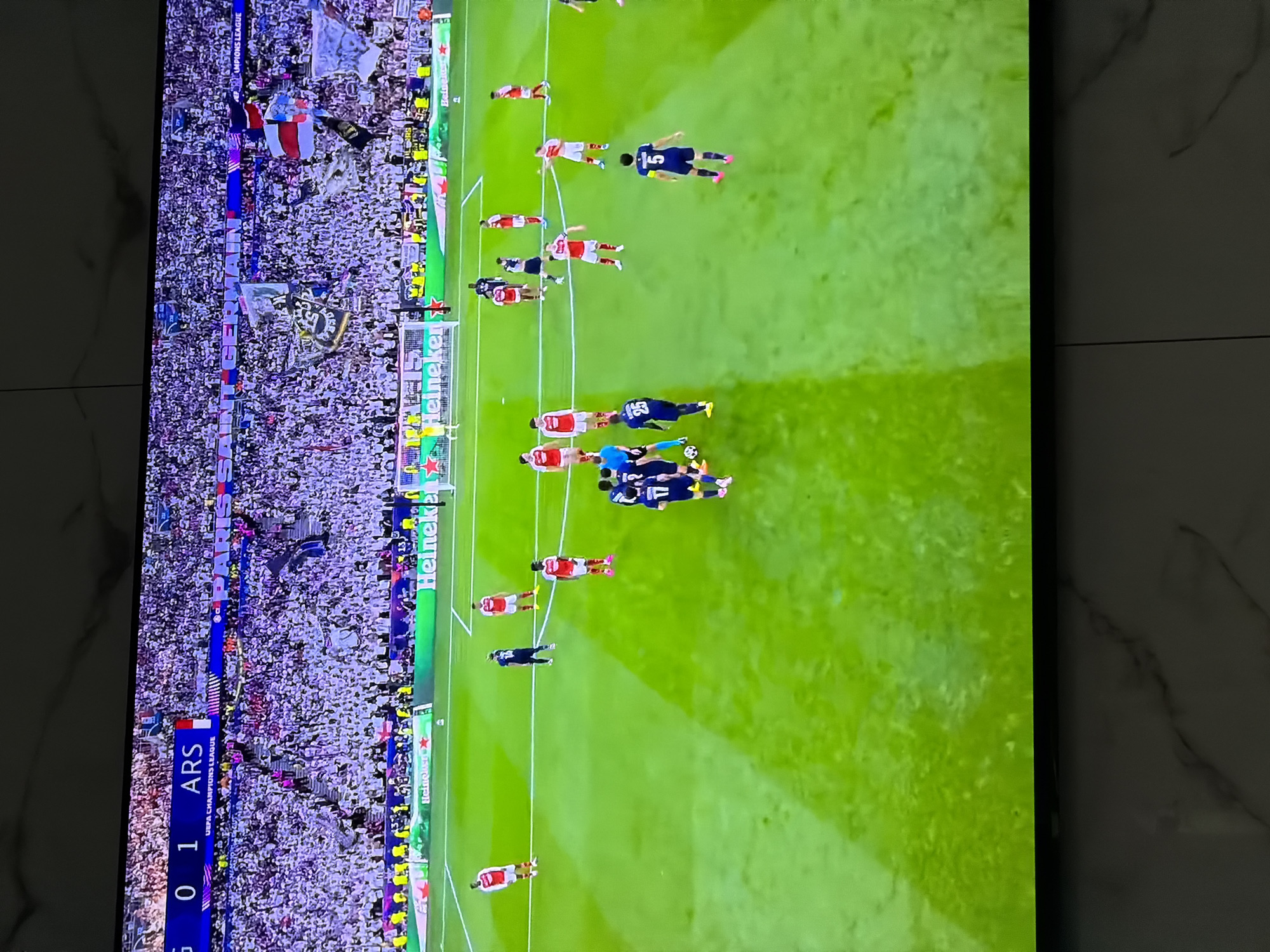 The image shows a screen displaying a soccer game between an unknown team and Arsenal (ARS), where Arsenal is winning 1-0. The game is part of the UEFA Champions League. There is a large crowd in the stands behind the goal, and the field of play shows players in both red and blue uniforms. There are Heineken advertisements along the edge of the field. The location, Asenovgrad, Bulgaria, is not visible in the image.