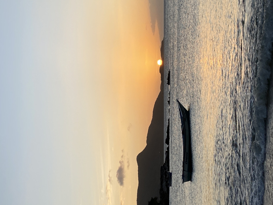 A tranquil seascape captures a sunset over water. Silhouetted mountains and a few boats dot the horizon, one prominently in the foreground. The sun casts a golden glow on the sky and reflects on the ocean surface where gentle waves meet the shore. The sky transitions from a clear blue to the warm hues of sunset.