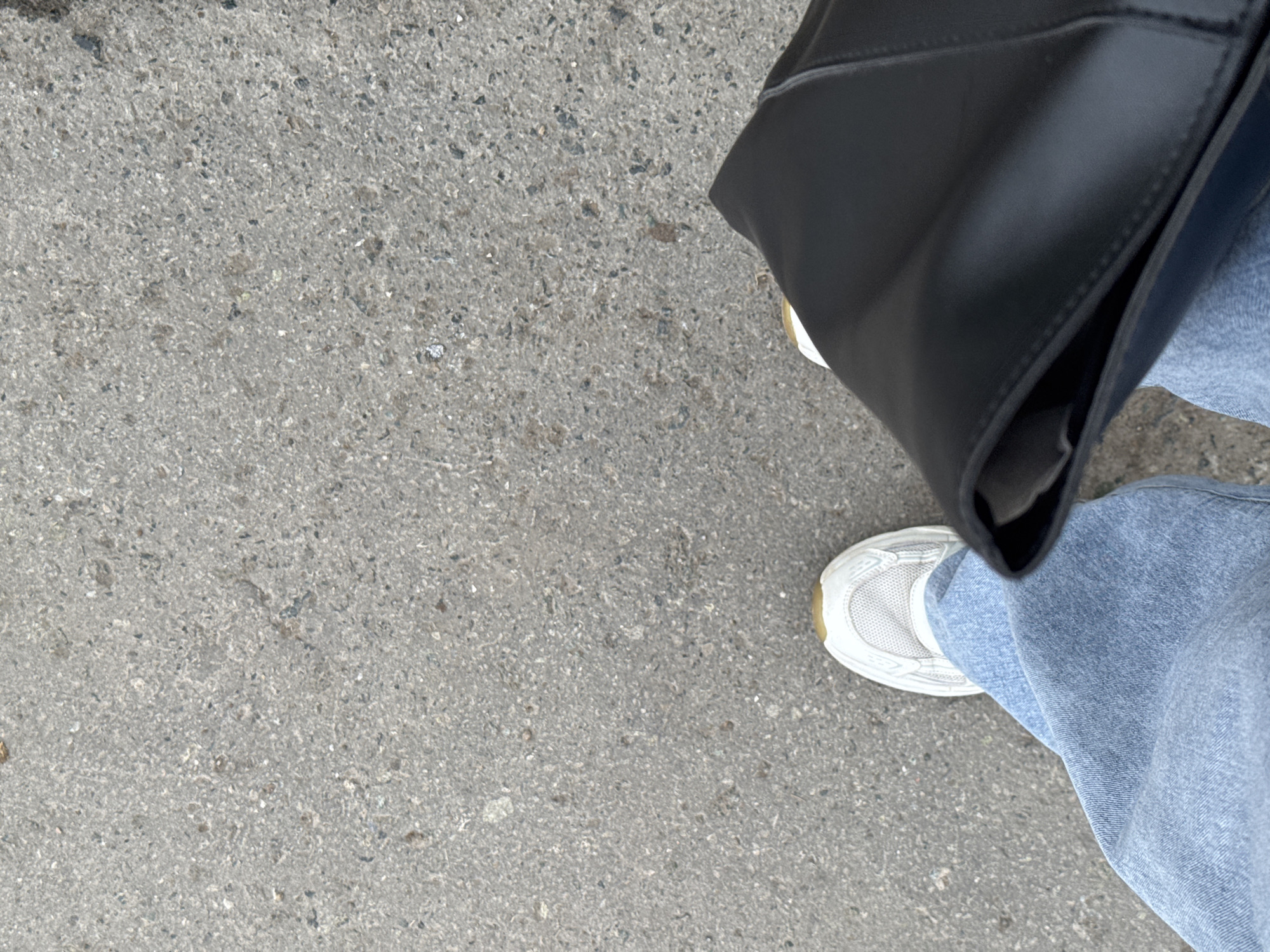 A first-person perspective shows a section of gray concrete pavement. A portion of a black leather bag is visible on the right, next to a person wearing light blue jeans and white sneakers. The location cannot be determined from this image.