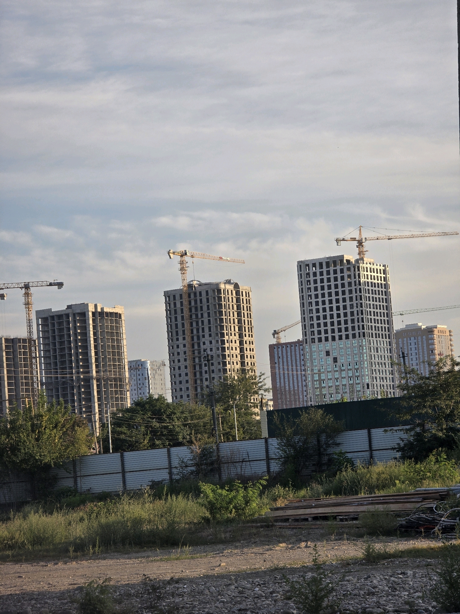 The image captures a construction site in the city of Grozny, Russia, featuring several unfinished high-rise buildings and multiple construction cranes. The sky is overcast with scattered clouds. In the foreground, there is a gravelly surface with some vegetation and a metal fence. Beyond the fence, the lower portions of the buildings are visible, some with exposed concrete structures and scaffolding, while others are partially clad in glass or facade panels. Construction cranes, predominantly yellow, are positioned around the buildings, indicating ongoing work. The overall scene suggests a developing urban area.
