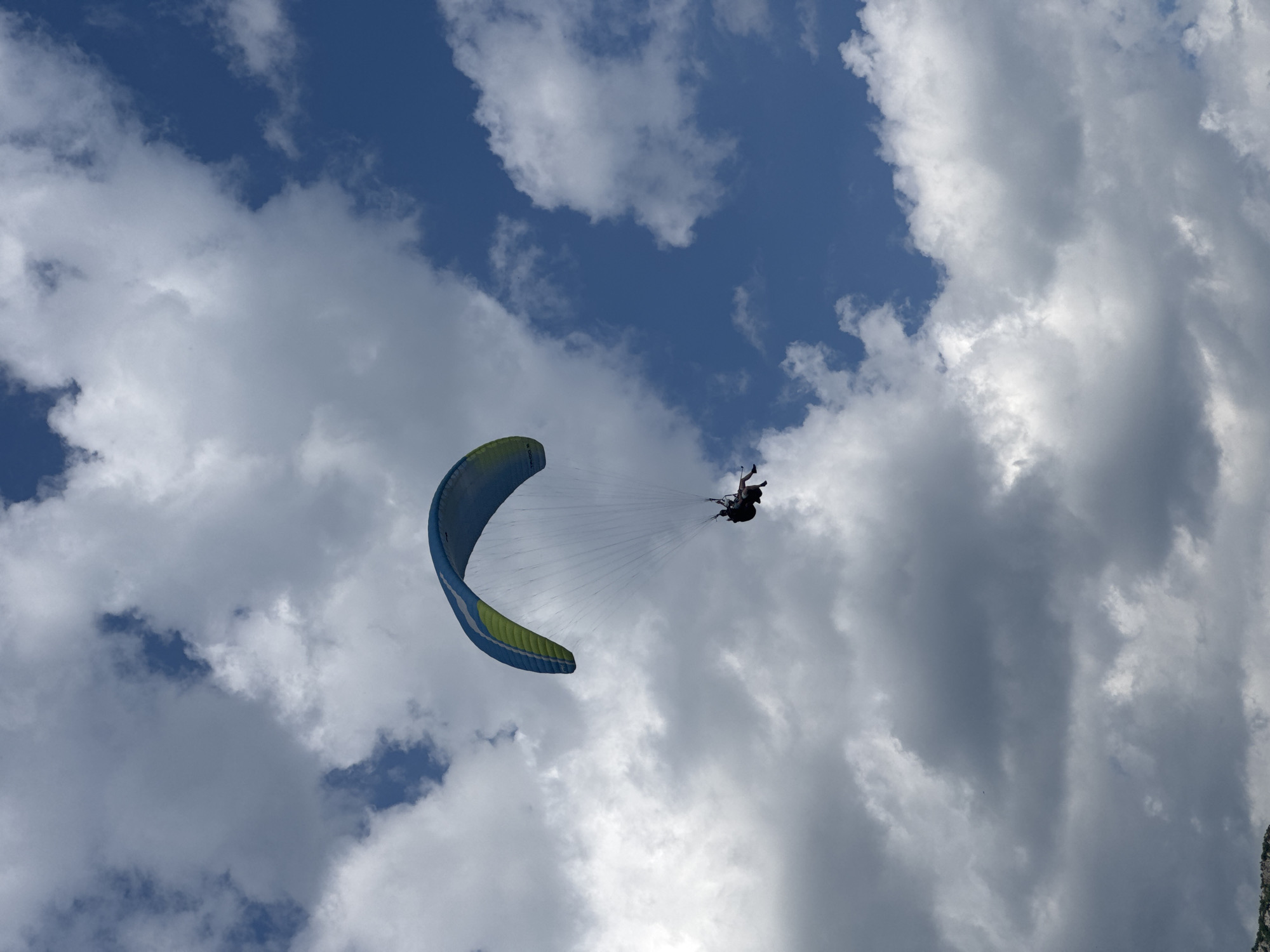 The image depicts a paraglider in flight against a backdrop of a partly cloudy sky. The sky transitions from a blue to a thick, white cloud cover. The paraglider is equipped with a blue and yellow wing. The pilot is harnessed below the wing, appearing to be in control of the paraglide. The location is Boreti, Montenegro.