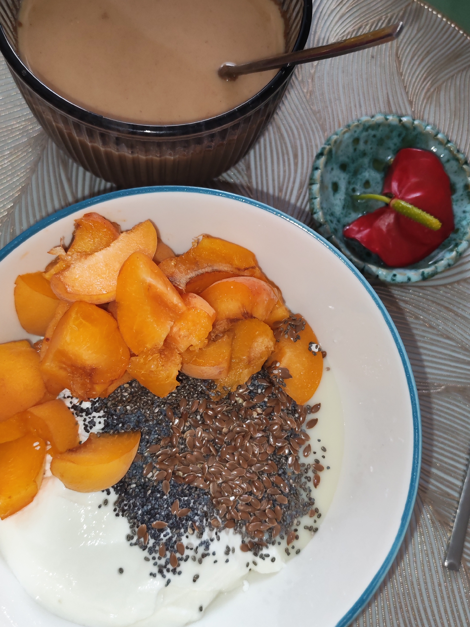 A top-down close-up shot reveals a white bowl filled with yogurt, topped with sliced peaches, chia seeds, and flax seeds. To the right, a small turquoise dish holds a vibrant red anthurium flower. In the background, a large glass bowl contains a creamy brown liquid, possibly a smoothie or soup, with a spoon resting inside. The setting appears to be a home dining table in Izhevsk, Russia, indicated by the presence of these food items and floral decoration, common in domestic settings. No individuals are visible in the frame.