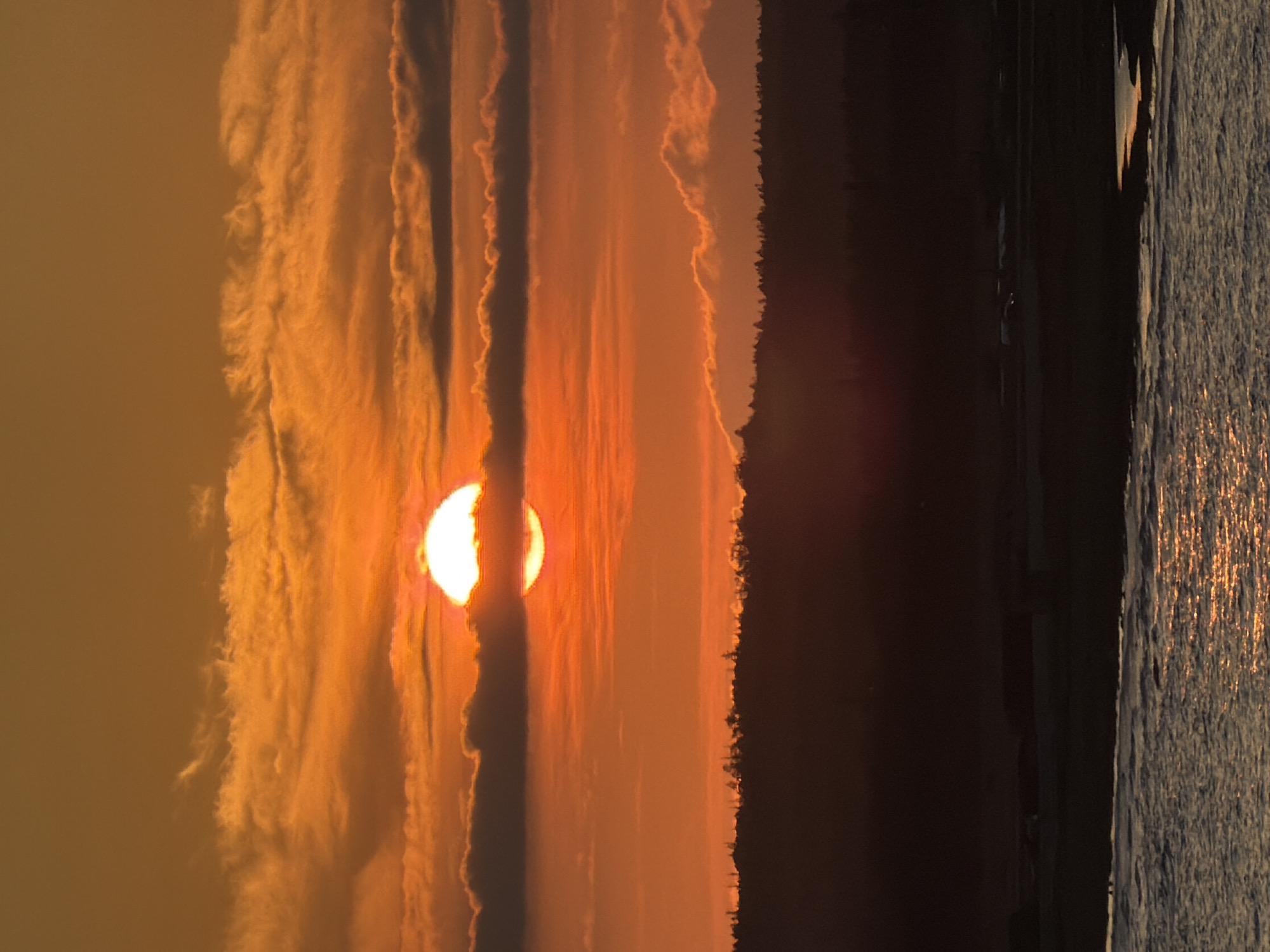 This is a photograph of a sunset over the ocean, taken from a position that overlooks a coastal landscape. The sun, a bright orange orb, is partially obscured by dark, textured clouds in the middle ground. The sky above is a gradient of orange and yellow hues, with wispy clouds catching the light. In the foreground, the water reflects the warm colors of the sunset, creating a shimmering surface. The middle ground features a silhouetted treeline, suggesting a forested area bordering the water. Beyond the treeline, the landscape is dark and indistinct, with subtle hints of structures or vehicles visible. The overall atmosphere is one of natural beauty and tranquility at the close of day.
