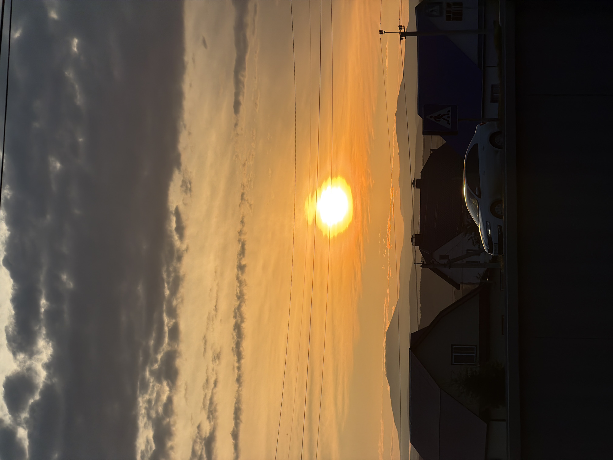 The image depicts a sunset scene with a bright sun radiating a warm orange glow through a hazy sky. Wispy clouds are visible, with some darker, heavier clouds on the left side of the frame, suggesting a mix of clear and overcast conditions. Power lines stretch across the sky, adding a linear element to the composition. In the foreground and mid-ground, silhouetted houses with pitched roofs are visible, indicating a residential or semi-rural setting. A white car is parked on the right side of the image, partially obscured by shadow. A pedestrian crossing sign with a blue background and white markings is also visible on a pole. The overall mood is serene, with the warm colors of the sunset creating a peaceful ambiance. The time of day is clearly sunset, given the position and color of the sun. No people are visible in the image.