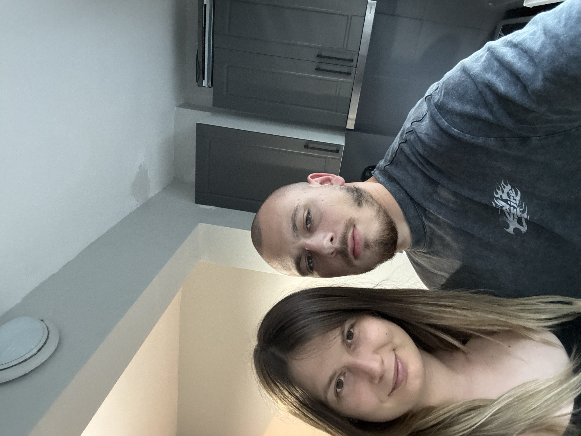 A selfie of a young man and woman is taken in a kitchen. The woman is on the left, her face turned towards the camera, and her hair is light brown and styled in long waves. She is wearing a black tank top. The man is on the right, his head shaved, and he has a goatee and mustache. He is wearing a dark grey t-shirt. Behind them, dark grey kitchen cabinets are visible. The lighting in the room suggests it is daytime, with light coming from the left and a ceiling light fixture visible at the top left of the image. The location is identified as Stara Zagora, Bulgaria.