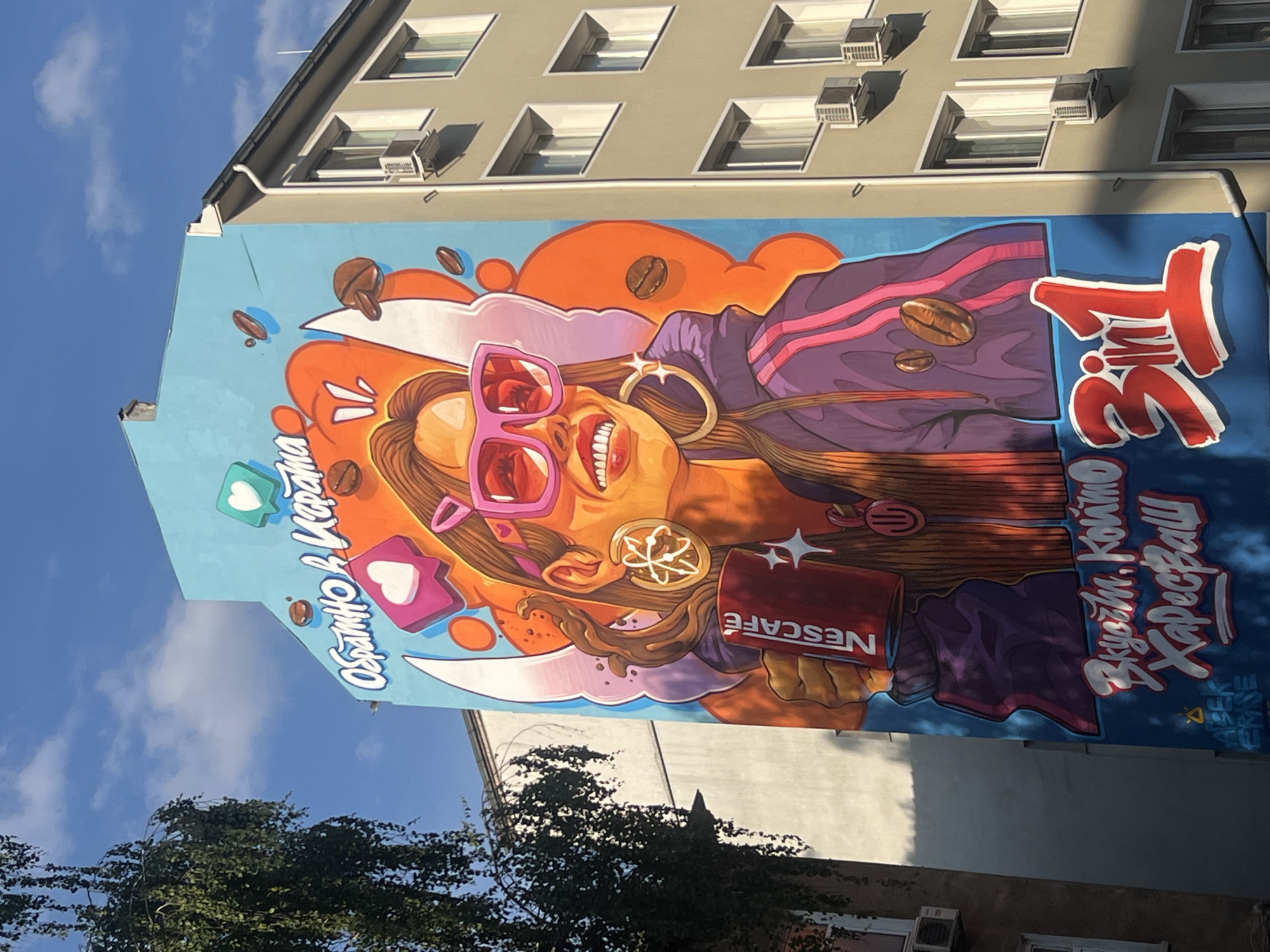 The image displays a large, vibrant mural painted on the side of a multi-story building in Sofia, Bulgaria, under a clear blue sky with sparse clouds. The mural, promoting Nescafe 3 in 1, features a stylized depiction of a young woman with long brown hair, wearing large pink reflective sunglasses and a purple track jacket with pink stripes. She has a wide, open-mouthed smile, revealing white teeth, and wears large gold hoop earrings; one earring has an atomic symbol design, and the other has stars. She holds a red mug branded "NESCAFE" in her left hand. Behind her head are stylized white wings, set against a dynamic background of orange, purple, and blue shapes, interspersed with floating coffee beans and two social media "like" bubble icons containing white hearts.
The mural includes several lines of text in Cyrillic. At the top, in white script, it reads "Обратно в Играта" (Back in the Game). Towards the bottom, in white script, it reads "Вкусове, които Харесваш" (Flavors you like), accompanied by a large red "3 in 1" logo. A small, indistinct signature or tag, possibly "A.BEK.VERSE", is visible in blue at the bottom left corner of the mural.
To the right of the mural, an adjacent multi-story building, light grey or beige in color, is visible. It features multiple rectangular windows and numerous white external air conditioning units mounted beneath most windows. The roofline includes a visible rain gutter. In the upper left quadrant of the image, the dense, drooping green foliage of a tree partially obscures the mural building and the sky. Strong sunlight illuminates the scene, casting distinct shadows on the adjacent building and the lower portions of the mural.