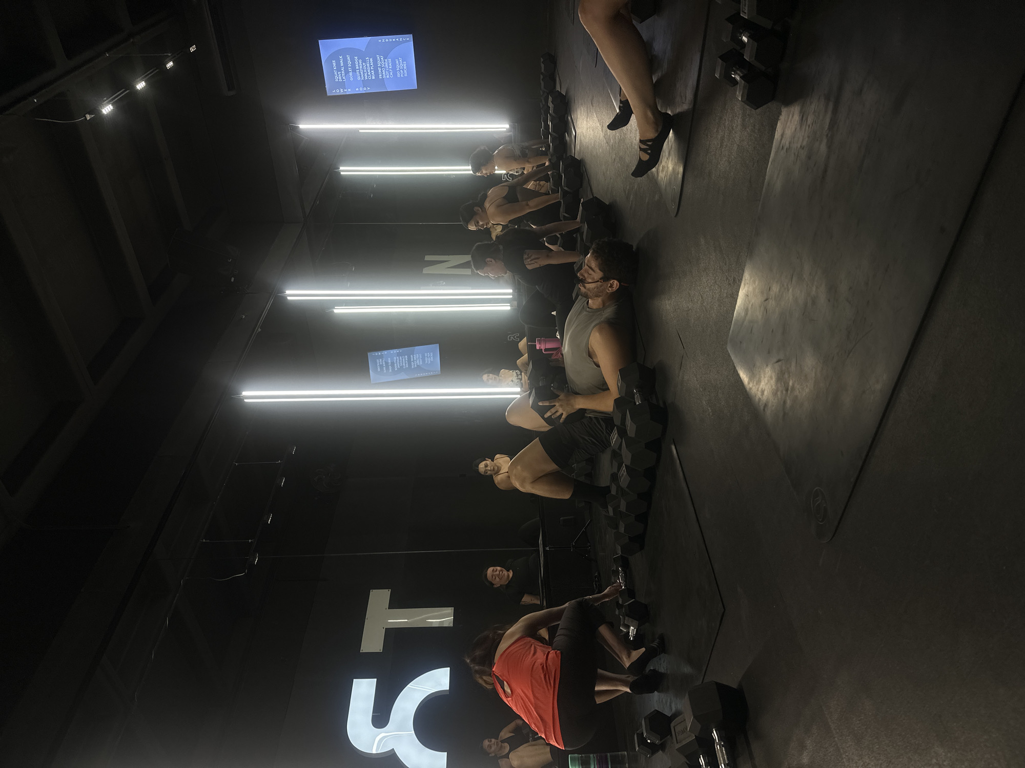 This photograph depicts a fitness class in progress within a modern gym or fitness studio. A line of individuals is seen exercising on mats, each using dumbbells of varying sizes. The room is illuminated by several linear LED light fixtures mounted on the ceiling, which creates a stark contrast against the dark walls. A screen displaying workout instructions is visible in the background. The gym's branding is incorporated into the decor with large illuminated letters "STNR" on the wall. The exercise mats are laid out in rows, and the floor appears to be made of a dark, durable material suitable for workout activities. No specific location within the city of Puebla, Mexico is identified in the picture.