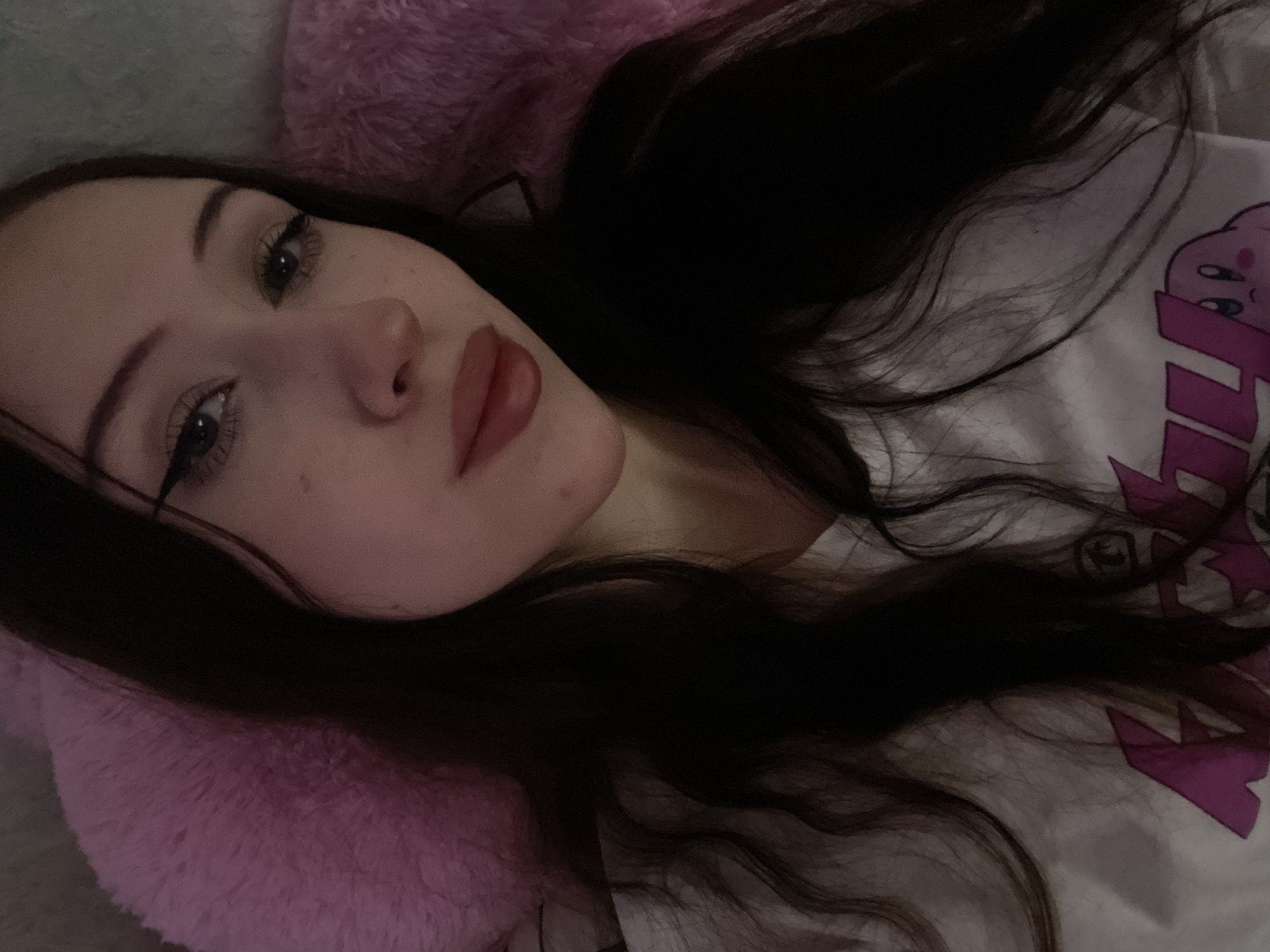 The image shows a close-up of a young woman with long, dark hair. She is wearing a white t-shirt with a pink design featuring the character Kirby. She is lying down, leaning against a fluffy pink pillow. The lighting is dim, suggesting an indoor setting. The woman has on eyeliner and lipstick. There is no information in the image to determine her location in Votkinsk, Russia.