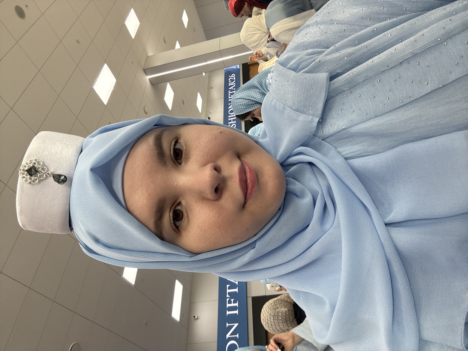 A young woman with light skin and brown eyes faces the camera, displaying a subtle smile. She wears a light blue hijab and a matching garment adorned with small, reflective embellishments. A small, rectangular white cap, embellished with a silver-colored pendant featuring a dark gemstone, rests on top of her hijab. In the background, several individuals, mostly women wearing various colored head coverings (including light blue, beige, white, and red), are visible. The indoor setting features a high ceiling with white square acoustic tiles and recessed square lighting fixtures. A dark blue sign with white lettering displaying "ON IFTAR" and "FASHION IFTAR26" is prominent in the mid-ground. A security camera is mounted on a background wall. The scene is located in Bol'shie Kabany, Russia.
