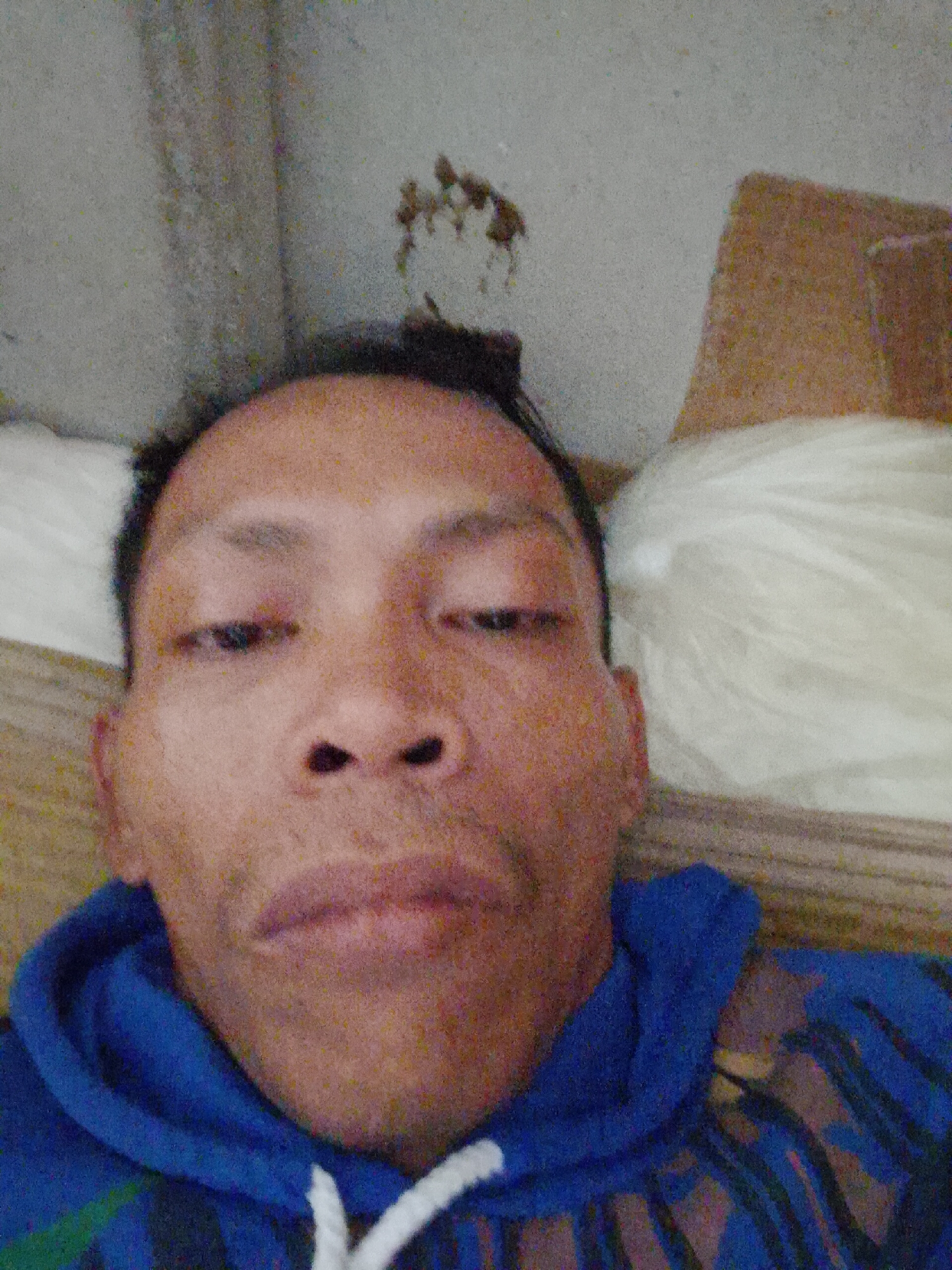The image shows a person with dark hair and brown skin. The person is wearing a blue hoodie with white drawstrings. The background includes a white pillow, a tan object (possibly cardboard), and a wall with some dark marks on it. The location is Bukit Ketok, Indonesia. The person appears to be lying down, possibly taking a selfie. The lighting is poor, resulting in a grainy image.