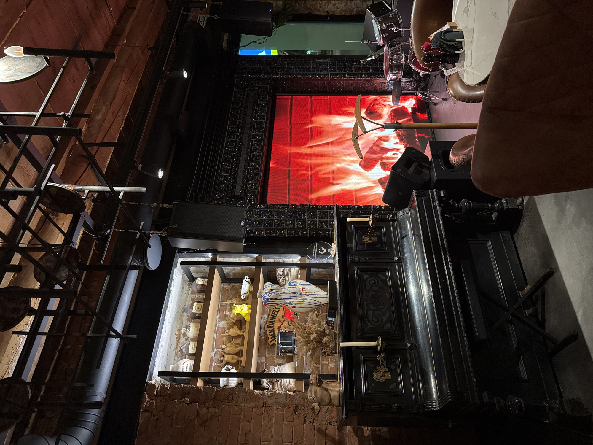 The image depicts an interior setting in Unknown, Unknown, featuring a blend of industrial and decorative elements. On the left, there's a structural steel framework with suspended circular lighting fixtures, juxtaposed against a brick wall. Adjacent to this, a shelving unit houses an assortment of items, including liquor bottles, decorative objects, and a "Jack Daniel's" sign.
The backdrop includes a large display screen showing a fiery animation, with mining tools positioned in front of it. A drum set is visible in the upper portion of the scene. To the right, an ornately decorated black cabinet stands with candlestick holders attached to it. Furniture is partially visible, including a tan chair and a white marble table.
The overall impression is of a bar or restaurant with a unique, thematic interior design. The lighting is subdued, emphasizing the screen display and decorative elements.