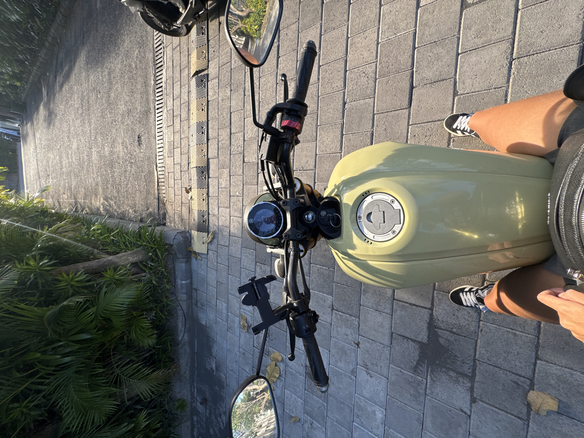 A high-angle shot depicts the front portion of a light yellow motorcycle parked on a paved surface, possibly a sidewalk or pedestrian area. The motorcycle features black handlebars with mirrors, a speedometer, and a tank cap. The background includes a road, some vegetation, and what appears to be drainage infrastructure.
A person wearing black and white sneakers, gray shorts, and potentially a helmet is partially visible on the right side of the frame, implying they are seated on the motorcycle. The photograph was captured during daylight hours, with sunlight casting shadows on the pavement.
The location is determined to be in North Kuta, Indonesia. Specific geographic references (e.g., street names, landmarks) are absent.