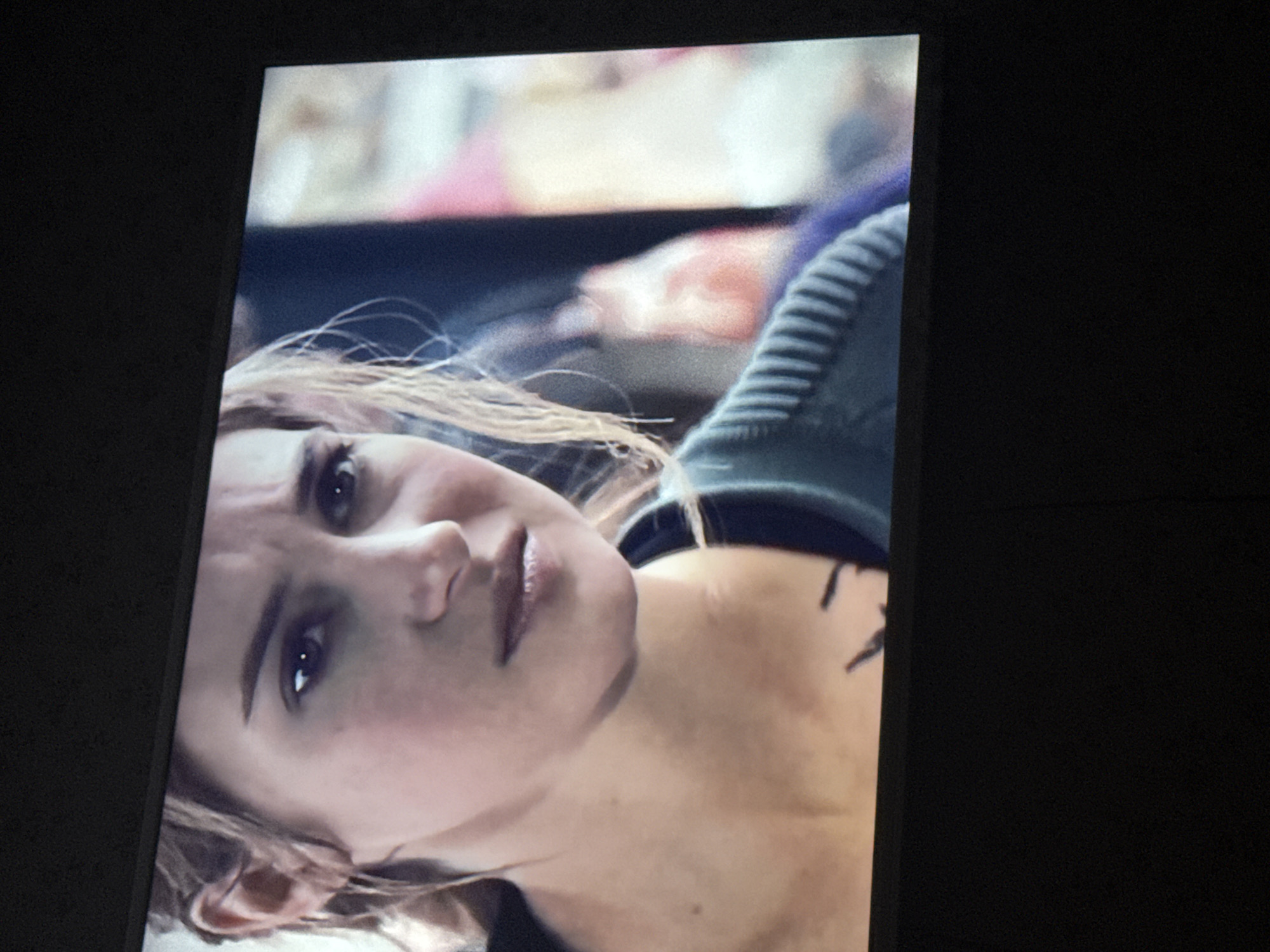 The image shows a close-up of a woman's face on a screen, possibly a television or computer monitor, set against a dark background. The woman has light skin, light brown hair, and is looking upwards with a slightly concerned expression. She has dark eyebrows and appears to be wearing some makeup. Parts of her clothing, including a dark shirt and a grey, ribbed shoulder, are visible. A small tattoo is seen on her neck. The location, Mytishchi, Russia, is not apparent from the visual information provided.