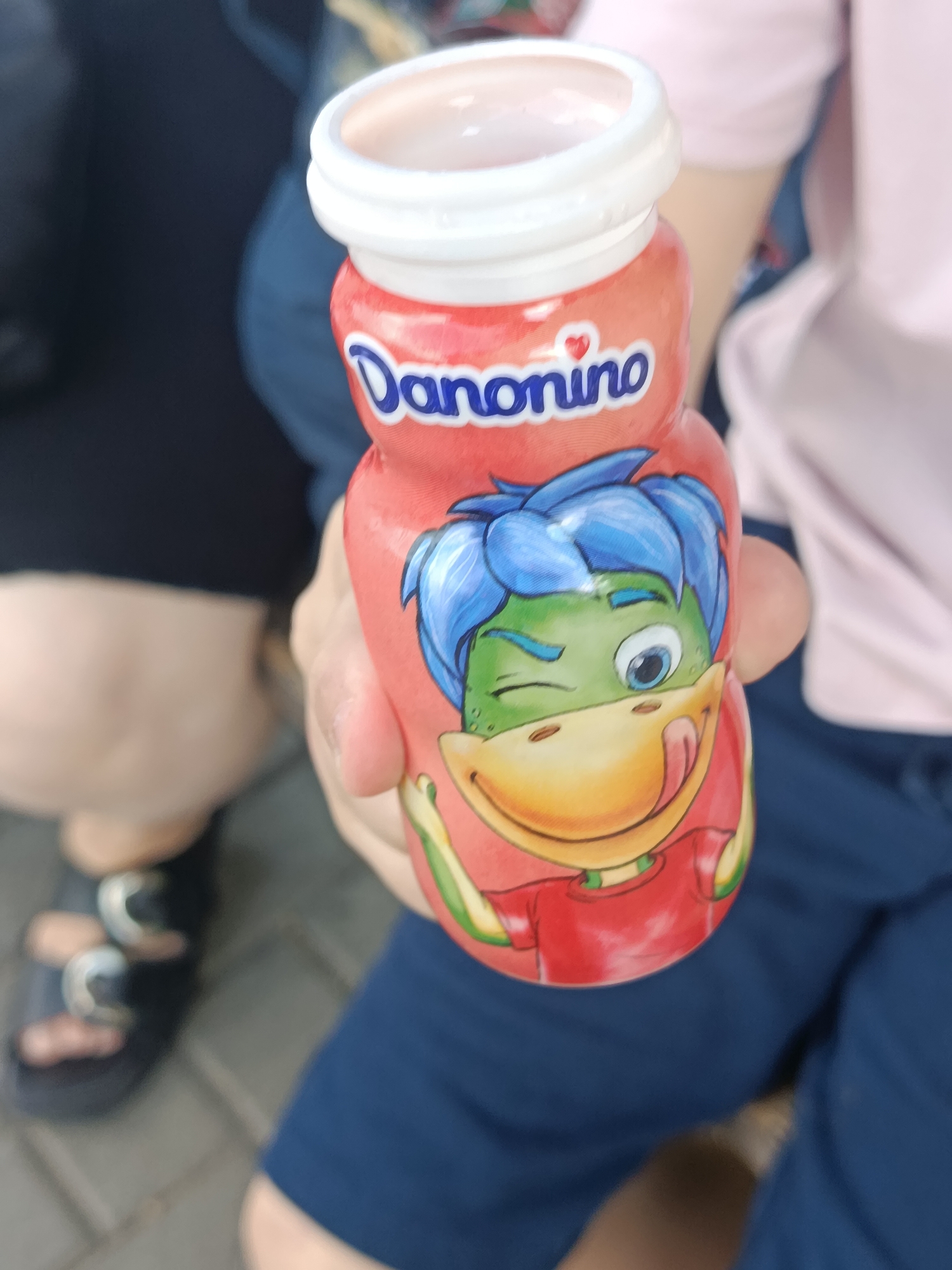 A person's hand is holding a red Danonino yogurt bottle featuring a cartoon character with green skin, blue hair, and yellow beak. The character is winking and sticking its tongue out. The bottle has a white cap and blue text that reads "Danonino" with a red heart above the "i". The person's dark clothing is visible on the left side of the image, and dark blue shorts are visible on the right. In the background, a blurred figure wearing a pink shirt and dark blue shorts can be seen. The ground appears to be paved with light gray tiles. The image appears to have been taken outdoors on a sunny day.