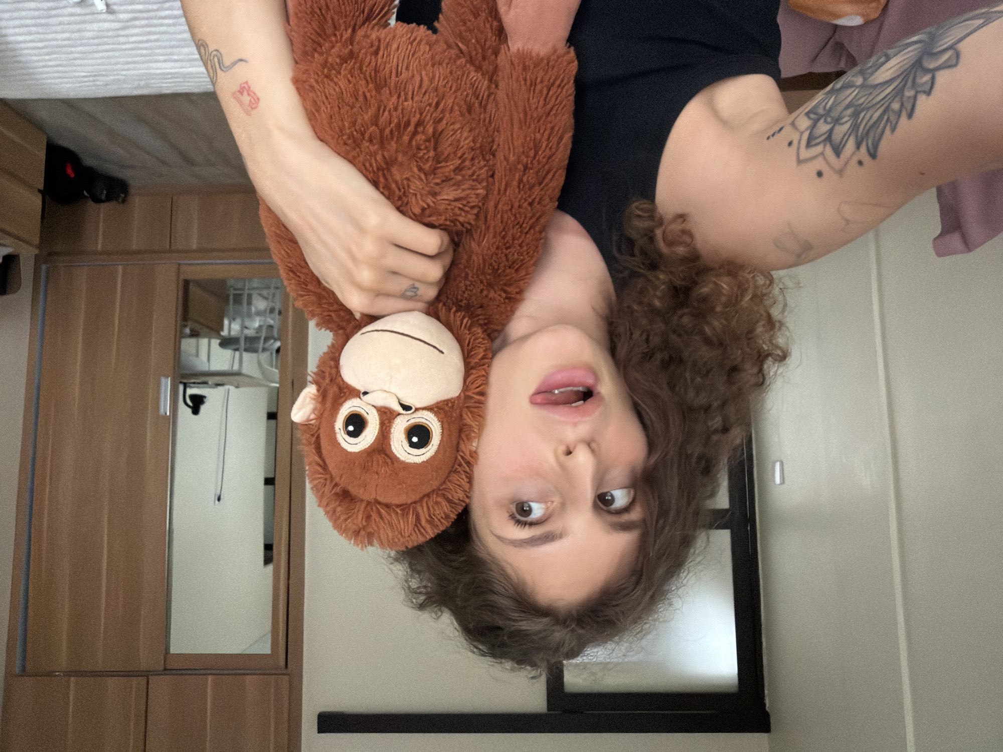 A young adult with curly brown hair, wearing a black tank top, holds a plush monkey toy. She is making a funny face, with her eyes looking to the side and her tongue sticking out. Her left arm, visible in the foreground, has a large tattoo of a flower. The background suggests an indoor setting, possibly a bedroom, with a wooden wardrobe and a door with a sliding glass panel. The overall scene appears casual and playful. The location is described as North Kuta, Indonesia.