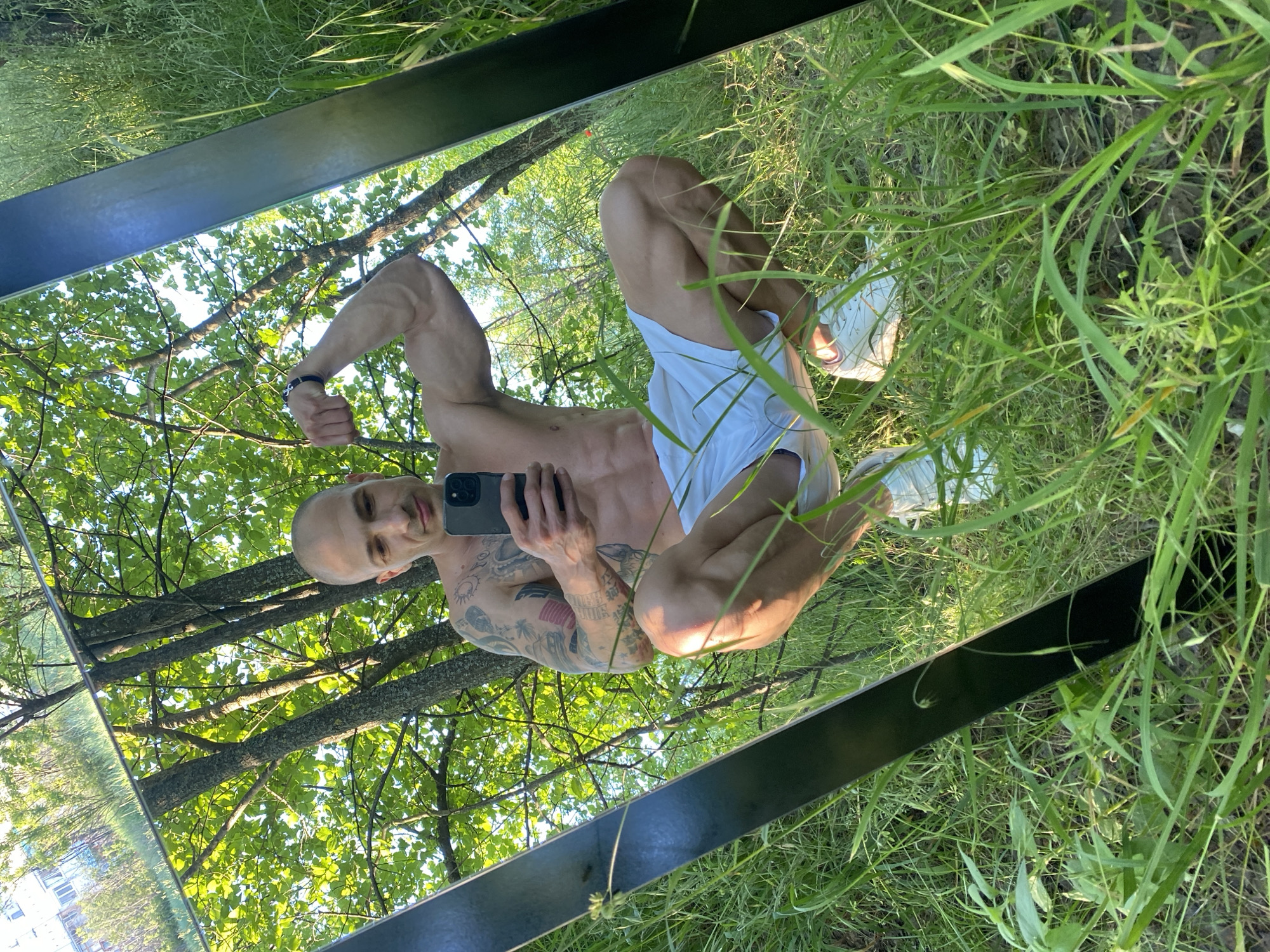 A bald, muscular man is reflected in a rectangular mirror set in tall grass in a wooded area. He is holding a black cell phone in his right hand, taking a selfie, and flexing his left arm. He is wearing white shorts and white sneakers. The background shows green trees with sunlight filtering through the leaves. Based on the surrounding trees and grass, the location could be a park in Pleven, Bulgaria.