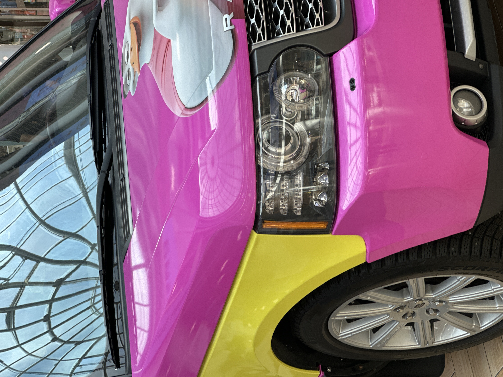 The image shows the front corner of a bright pink and yellow Range Rover. A cartoon duck graphic is visible on the hood. The vehicle is parked indoors, with a glass ceiling structure reflected on the windshield. The setting appears to be a modern shopping mall or exhibition space. There are no people visible in the frame. The car's headlight and grille are prominent. The license plate is obscured. No information about Minsk, Belarus is present in the image.