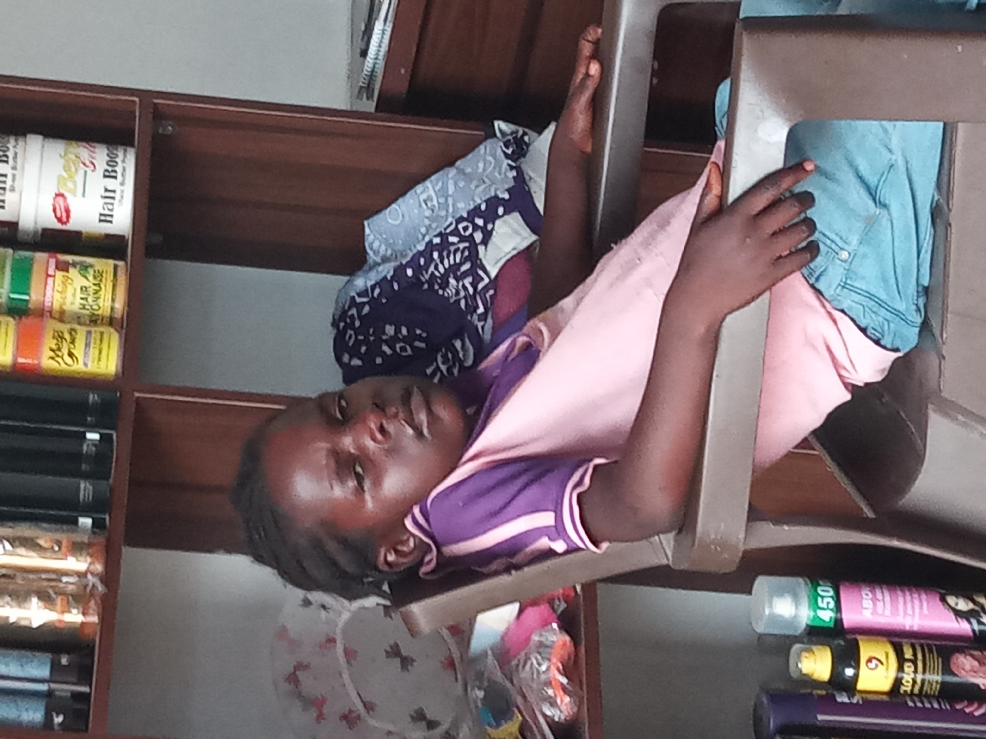 A young person with dark skin and dark hair is captured in a close-up, lying on a brown chair and looking directly at the camera with a neutral expression. She is wearing a short-sleeved shirt that is pink on the upper half and purple on the lower half, with white stripes on the sleeves. Her left hand rests on her stomach, while her right arm is extended behind her head, leaning against the chair frame.
The setting appears to be an indoor space, possibly a hair salon or a retail shop, indicated by the wooden shelves lining the background, filled with various hair care products. On the left side of the image, multiple jars are visible on a shelf; text like "Hair Boo" and "Hair Mayonnaise" can be clearly read on some of these containers. Other partially visible brands include "Mofajang."
Behind the person's head, a folded stack of patterned fabrics in shades of purple, white, and gray rests on the chair. A light blue garment, resembling jeans, is draped over the chair behind her body. On the right side and below the person, another shelf holds several spray bottles and cans. A prominent green and pink bottle displays "450," while a yellow and black bottle shows the number "9." The lighting is consistent with an indoor environment. This image was taken in Sabon Tasha, Nigeria.