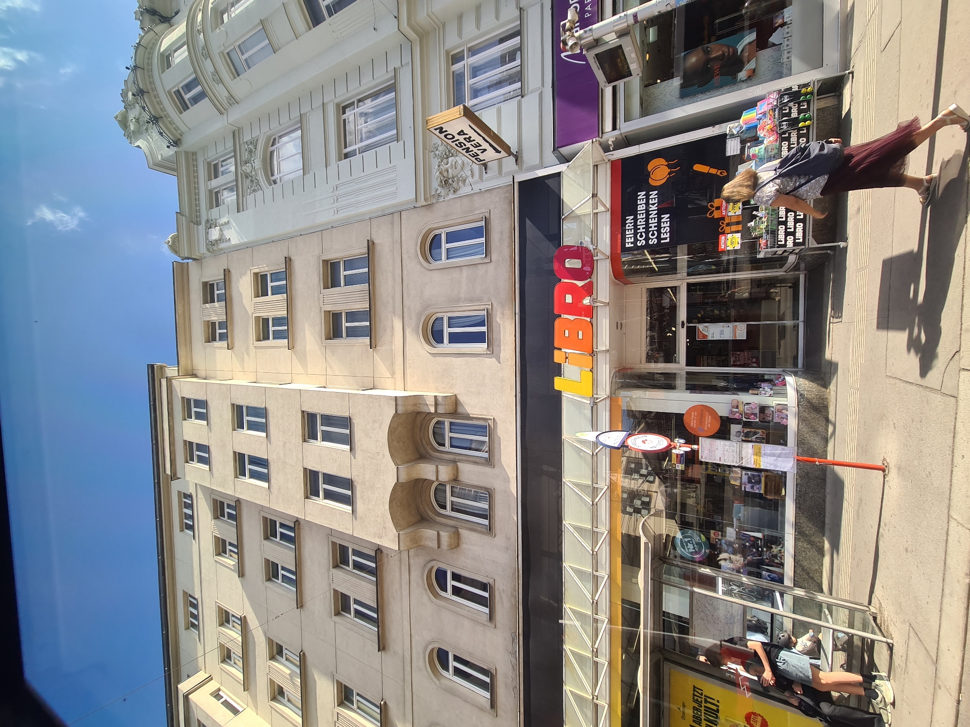 A low-angle, full-frame outdoor shot captures a street scene in Vienna, Austria, under a bright blue sky with sparse clouds. The buildings in the background are multi-story structures with cream-colored facades. On the left, a light yellow building with numerous rectangular windows dominates the scene. To its right, a more ornate pale green building features decorative architectural elements and a sign reading "PENSION VERA" with an arrow pointing to the right.
At street level, a bright orange and yellow "LIBRO" sign signifies a bookstore. The store's storefront is a mix of glass windows displaying merchandise and a prominent black banner with German text: "FEIERN SCHREIBEN SCHENKEN LESEN." This translates to "Celebrate Write Gift Read." In front of the store, a display rack holds various books and other items.
On the left side of the image, in front of a glass-enclosed bus stop, two individuals are visible. One person, wearing a dark top and denim shorts, sits on the steps of the bus stop, holding a small stuffed animal. Another person, dressed in black, is partially visible behind them. To the right of the bookstore, a person with light brown hair wearing a patterned top and a long, dark red skirt walks away from the camera, carrying a dark blue bag over their shoulder. The sidewalk is paved with light-colored concrete.
