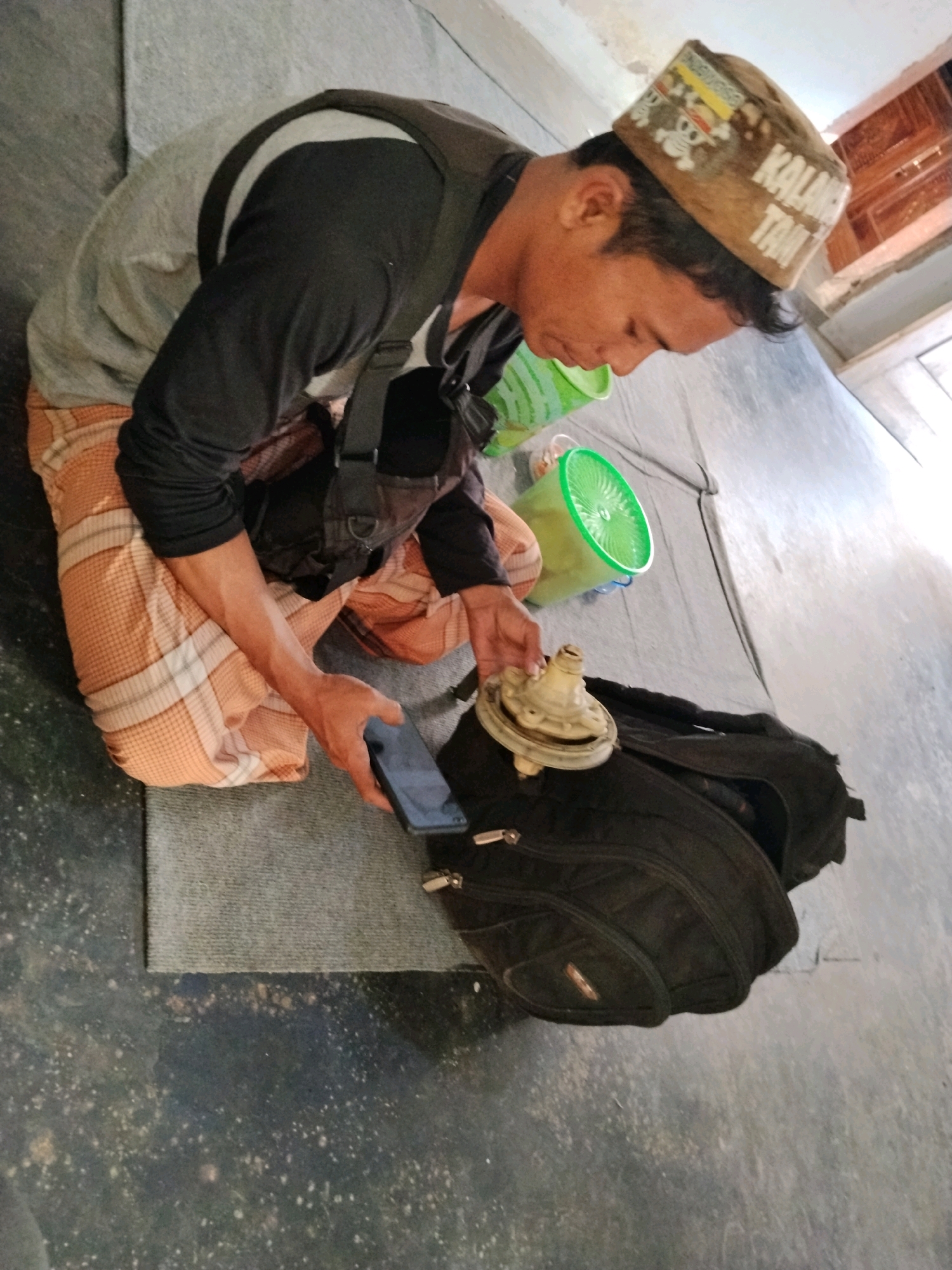 In Panggungan, Indonesia, a person wearing a tan and white checkered sarong, a grey long-sleeved shirt with black sleeves, and a brown cap with text is crouching on a textured grey mat. The person is holding a black smartphone in their right hand and reaching towards a white, dome-shaped object with a nozzle, which is resting on an open black backpack. To the left of the person, two green plastic buckets are visible. The ground is a dark, speckled surface.