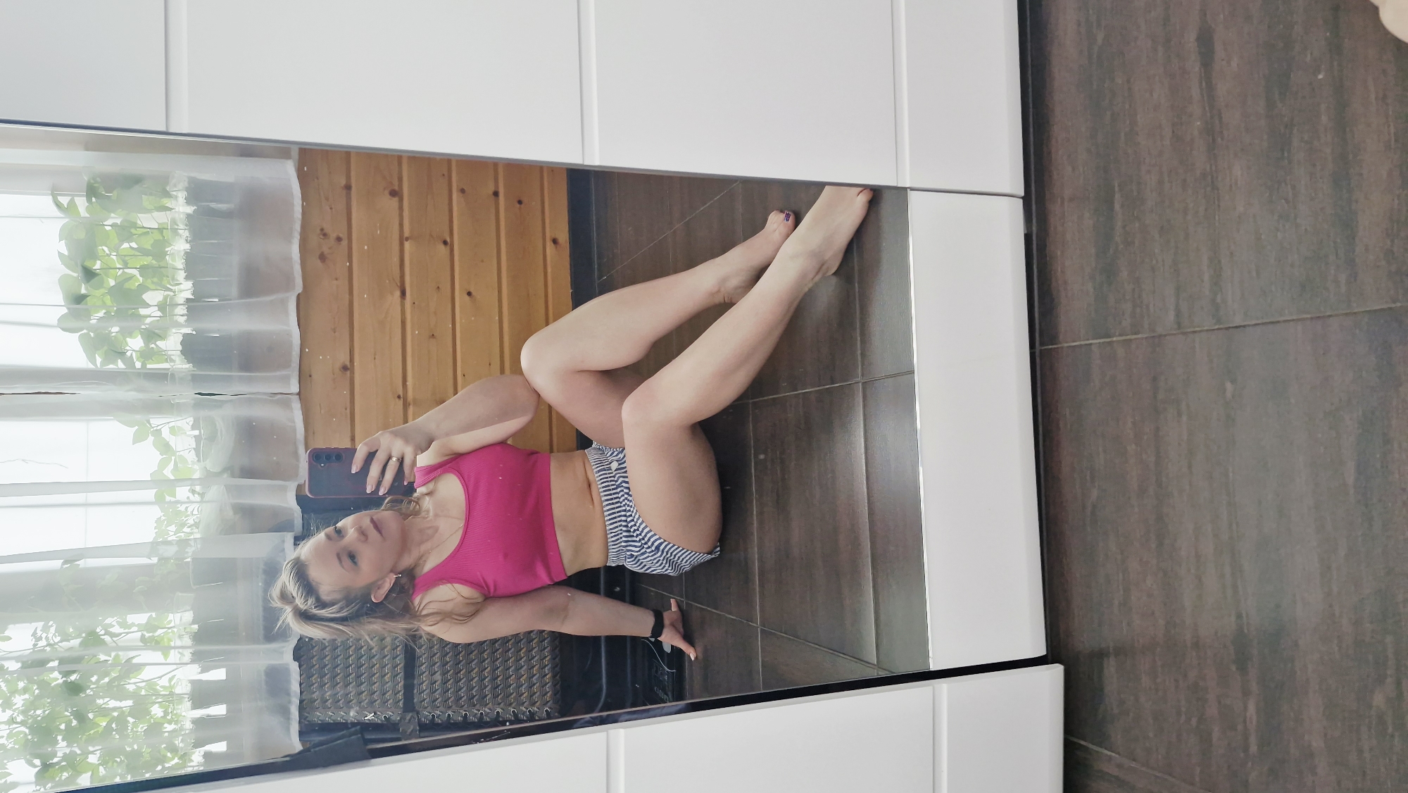 An adult woman is captured in a mirror selfie, lying down or reclining, with the image rotated 90 degrees clockwise. She has blonde hair and is wearing a bright pink sports bra or top and striped blue and white shorts. She holds a dark-colored smartphone in her left hand, and a ring is visible on her left ring finger. Her fingernails are light-colored, and her toenails are painted purple. A black wristband or watch is on her left wrist.
The mirror reflects an indoor setting, revealing a rustic wooden wall, a window with sheer white curtains, and small green plants behind the curtains. A piece of dark woven furniture is partially visible behind her. The floor in the reflection is dark, appearing to be tiled or dark wood. The foreground of the actual room, where the mirror is, shows white cabinetry or wardrobe panels. The room is brightly lit, likely by natural light from the window, indicating it's daytime. No visible text is present in the image. The location context provided is Otchor, Russia.