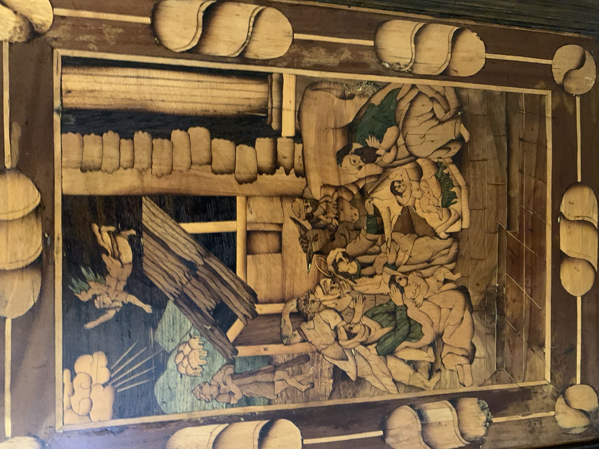 The image depicts a scene from the Nativity of Jesus, rendered in marquetry. The figures are composed of various wood tones. In the upper left, an angelic figure with wings descends from clouds, with rays of light emanating towards the scene below. A shepherd stands in the mid-ground to the left, holding a staff and looking towards a flock of sheep. The central focus is the stable, where Mary, Joseph, and other figures are gathered around the infant Jesus. Mary is depicted holding the baby, who is lying on a bed of straw. Joseph is seated beside them. Other figures, possibly shepherds or wise men, are present, some kneeling. To the right, there are architectural elements, including a column and a brick wall. The scene is framed by a decorative border of wood inlay. The style suggests it is an object, possibly furniture or a decorative panel, rather than a fresco or painting. There is no visual information in the image to associate it with the city of Poznan, Poland.