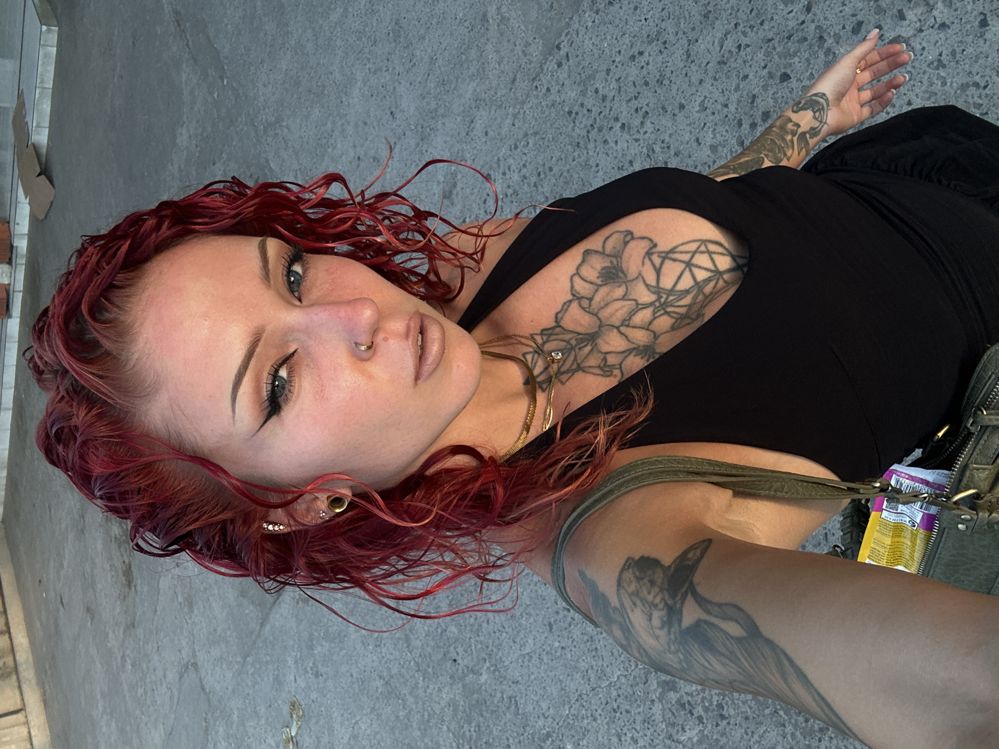 A woman with red curly hair, wearing a black top and multiple tattoos, takes a selfie. She has fair skin, light eyes, and a nose ring. Her tattoos are visible on her chest, upper arm, and forearm. She has ear piercings and wears necklaces. She is outside on a paved surface, likely concrete. There are buildings and potentially a sidewalk in the background. The location of Nusa Penida, Indonesia, is not confirmed by the image alone, but based on the location information the photo may have been taken there.