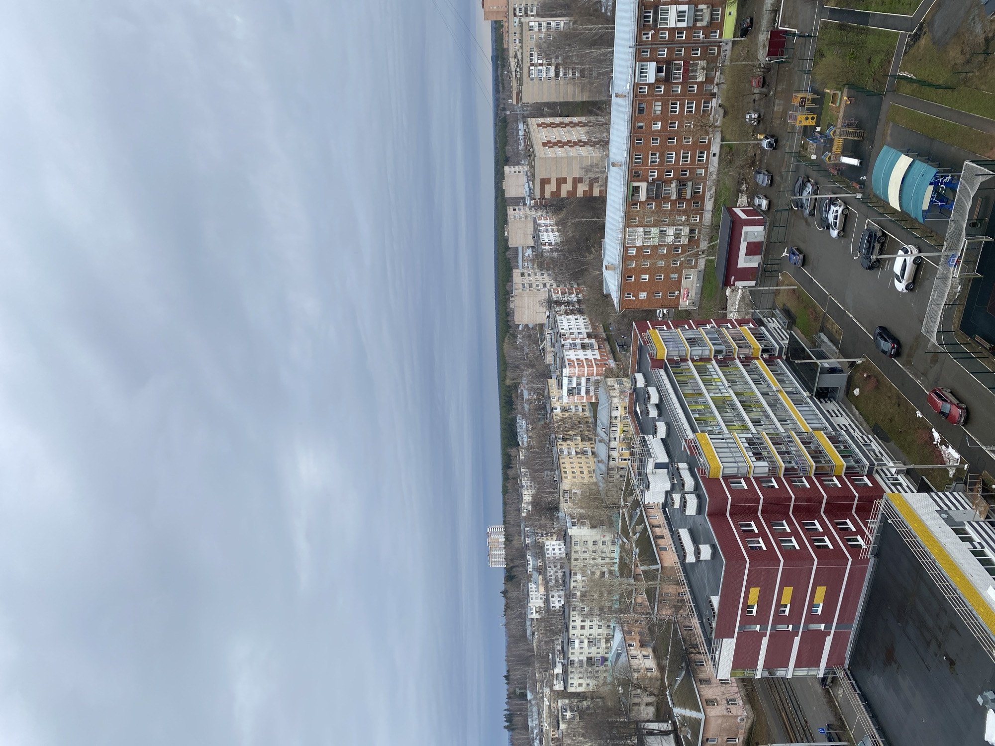 The image depicts a cityscape, likely taken from an elevated position. In the foreground is a building with red and yellow accents. Adjacent to it, there is a parking lot with several vehicles. Further in the distance, there are residential buildings interspersed with trees, leading to the horizon where the sky is overcast. The location is Izhevsk, Russia. There are no people immediately visible in the image.
