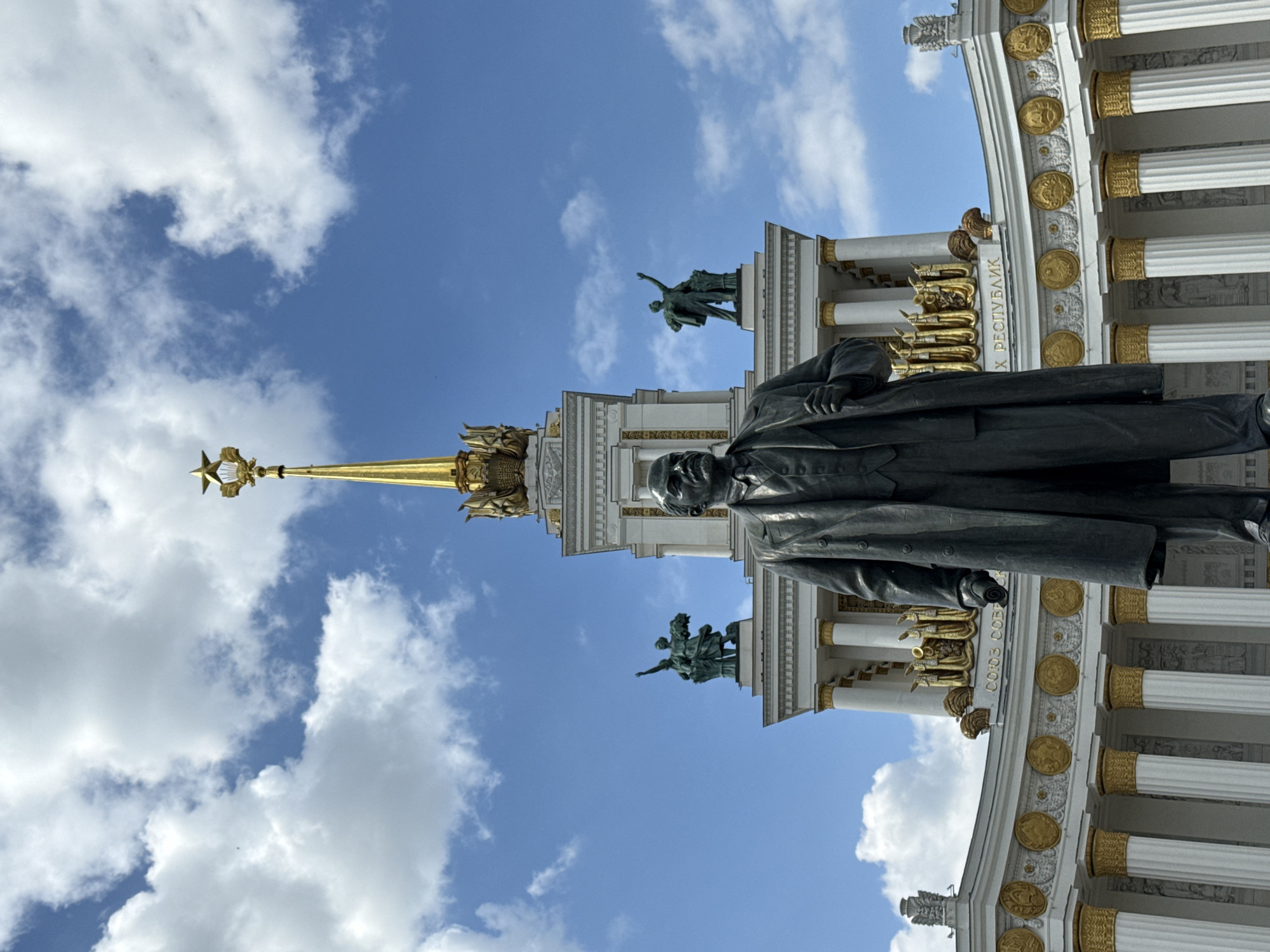 The image captures a scene at the Exhibition of Achievements of National Economy (VDNKh) in Moscow, Russia. The frame includes a bronze statue of Vladimir Lenin in the foreground, standing in front of the Central Pavilion of the VDNKh. The pavilion features white columns, ornate gold detailing, and sculptures on its roof. A spire topped with a gold star rises above the pavilion. The sky is blue with scattered white clouds.