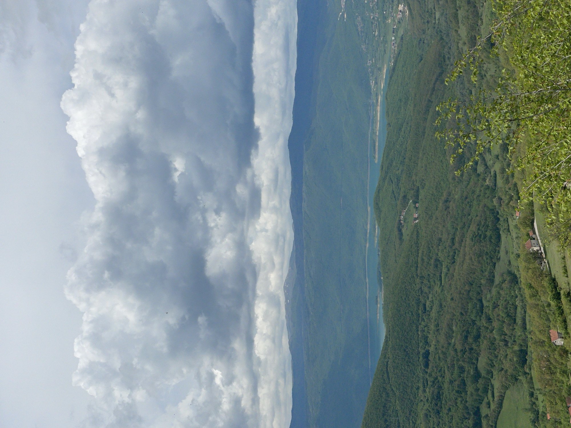The image shows a mountainous landscape in Gmići, Bosnia and Herzegovina. On the right, a lush green mountain slope covered in trees extends downwards. Some houses are sparsely scattered on the hillside. A body of water, likely a river or lake, snakes along the base of the mountain. In the distance, another, less defined mountain range fades into the horizon. Dominating the left side of the image are large, billowing clouds. A tree is in the top-right corner of the image.