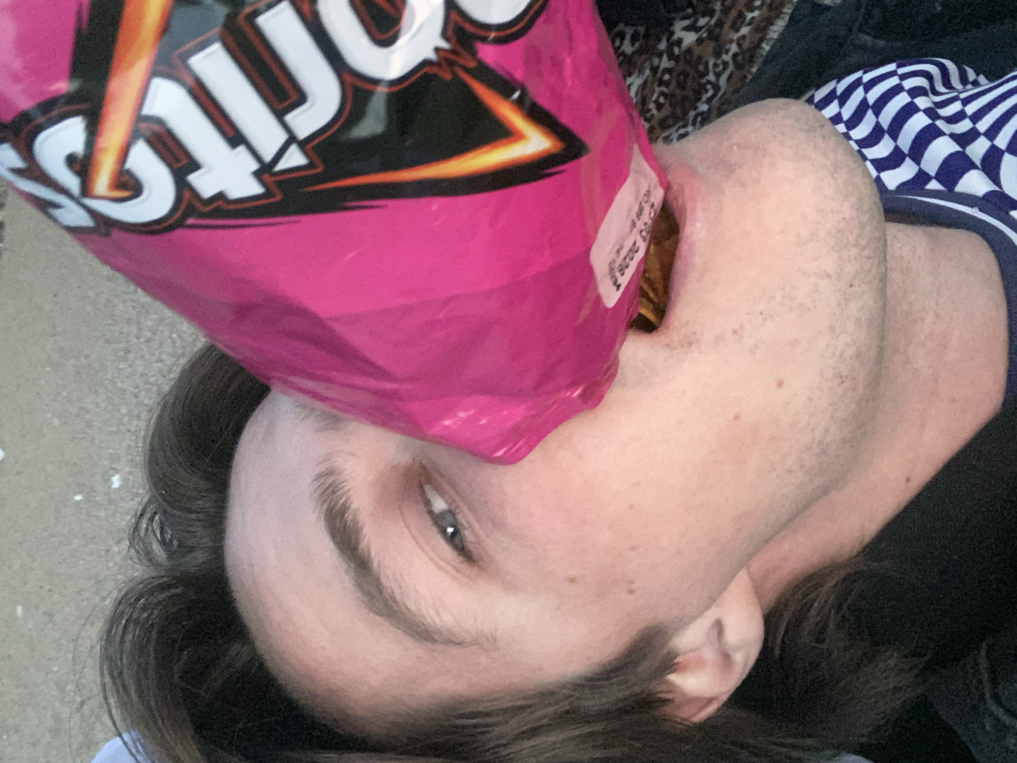 A person is lying down, facing the camera, with a pink bag of Doritos chips held over their mouth, pouring the contents into their mouth. The person has light skin, dark hair, and a blue eye visible. They are wearing a blue and white checkered shirt and a black shirt. The background is a light gray surface, and a corner of an animal print blanket is visible behind the person's head. The location is not discernible based on the given information.