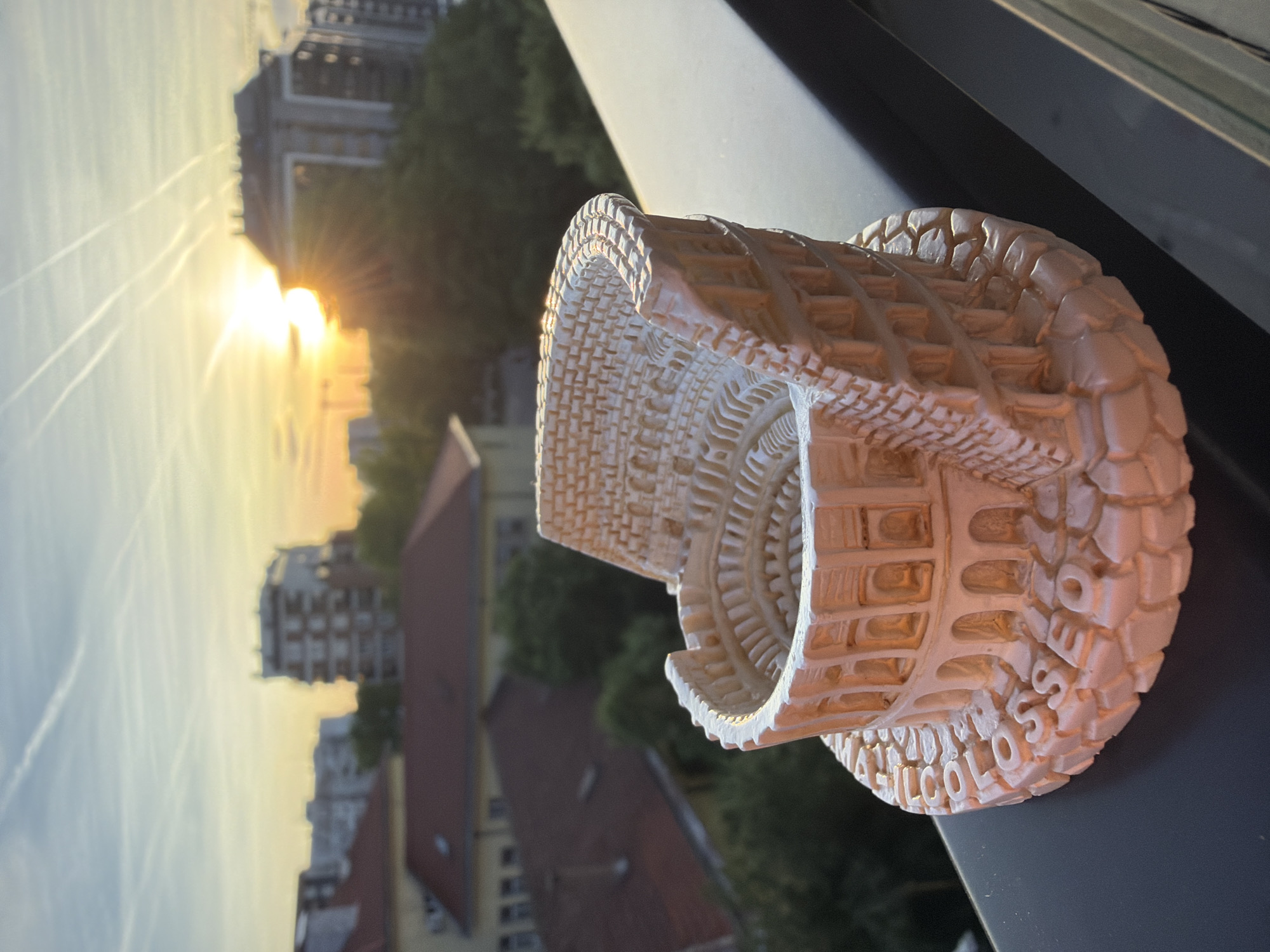 The image shows a miniature model of the Colosseum in Rome, Italy, placed on a surface. The model is detailed, showcasing the architectural features of the Colosseum. In the background, there is a cityscape with buildings, trees, and a sunset. The location appears to be a window or balcony overlooking the city. The model is partially illuminated by the sunlight.