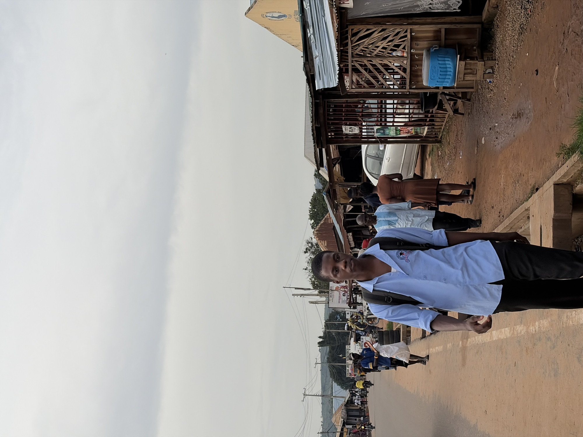 The image depicts a street scene in Sabojibi, Nigeria, under an overcast sky. In the foreground, a young man wearing a light blue collared shirt and dark pants walks towards the camera, carrying a black backpack. He appears to be the central subject of the photograph.
Along the street, other individuals are visible, some walking and others appearing to be stationary. To the right of the street, market stalls with corrugated metal roofs are set up, displaying various goods and merchandise. One stall has a wooden structure with a metal gate, and a blue cooler is prominently placed outside it. A white car is parked near this stall.
Further down the street, more market stalls, people, and vehicles, including motorcycles, are present, indicating a bustling commercial area. The road surface is made of dirt and appears to be uneven. The background shows a line of trees and buildings on a hillside, with power lines stretching across the frame, suggesting an urban or semi-urban environment. The overall atmosphere suggests a typical day in a Nigerian town.
