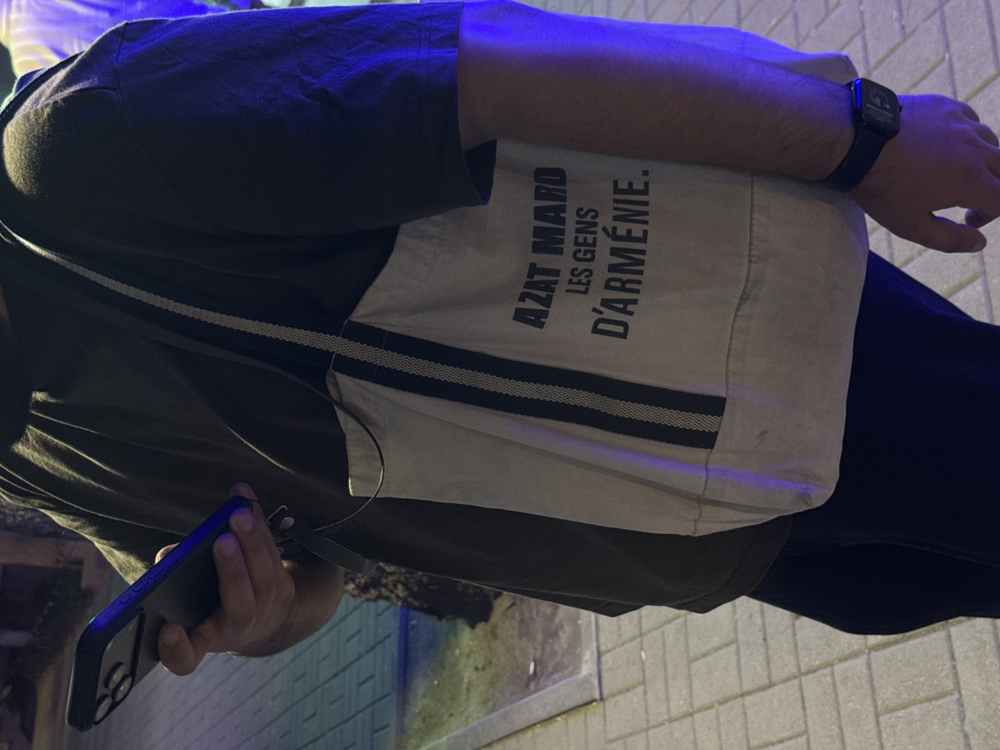 A person is depicted in an urban outdoor setting at night, likely in Yerevan, Armenia. The individual, observed from the chest down, wears a dark, possibly olive green, short-sleeved t-shirt and dark trousers or jeans. The right hand holds a dark-colored smartphone, consistent with an iPhone featuring a triple-lens camera system, enclosed in a dark case. The phone's side buttons exhibit a distinctive purple-blue illumination. A dark strap is attached to the phone case.
Slung over the left shoulder is a light-colored (off-white or beige) canvas tote bag, hanging on the right side of the person's body. The bag features two prominent dark vertical stripes and bold black text which reads: "AZAT MARD", followed by "LES GENS", and then "D'ARMÉNIE." This translates to "Free Man The People of Armenia." The person's left wrist wears a dark smartwatch with an illuminated blue display.
The ground is paved with rectangular light-grey pavers, indicative of a city sidewalk. Artificial lighting dominates the scene, casting strong blue and purple hues on the right side of the image, particularly on the person's arm and the tote bag. The background is blurred, showing hints of dark foliage or an object with a greenish tint on the left, and lighter, indistinct shapes with blue/purple light on the far right. The overall illumination suggests a nocturnal environment.