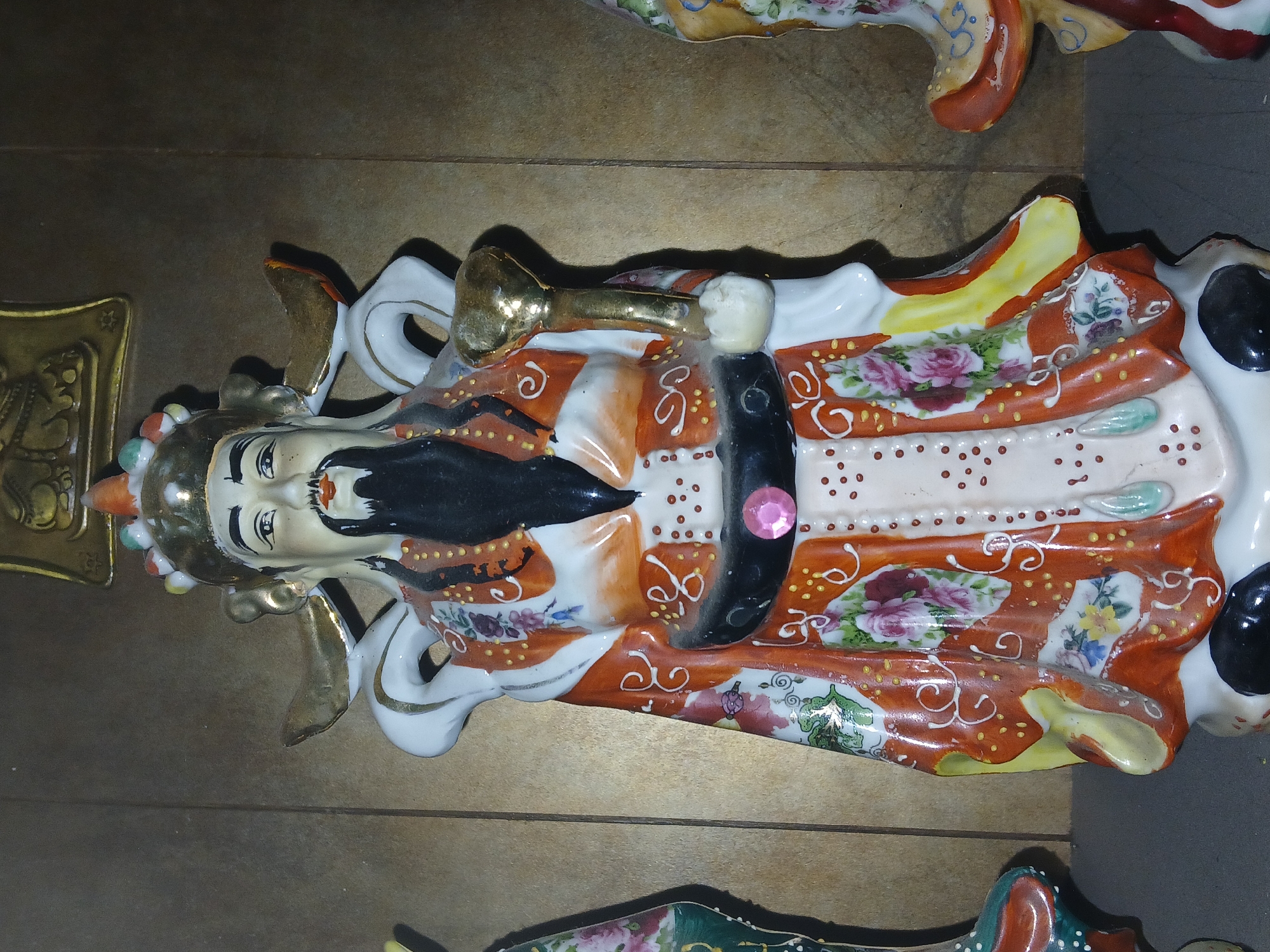 The image shows a decorative figurine of a man with a black beard, wearing an orange and white robe with floral patterns. The figurine is lying horizontally on a wooden surface. The man has a golden headdress adorned with colorful beads and is holding a golden object in his hand. The location is likely Bengaluru, India, though this is not discernible from the image. There are also parts of other figurines around.