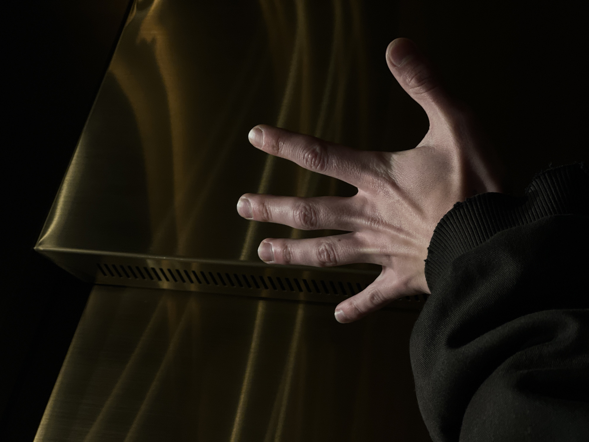 A human right hand is prominently displayed in the center of the frame, palm facing the viewer with fingers splayed. The hand appears to be that of an adult, with fair skin and visible, trimmed fingernails. A dark, possibly black, fabric sleeve with a ribbed cuff is visible at the lower left, indicating the garment worn by the individual. The background consists of dark, reflective metallic panels exhibiting a golden or brass coloration. These panels show subtle, undulating horizontal reflections of light and shadow. A vertical seam is present on the right side, where two metallic panels meet at an angle, featuring a strip of evenly spaced horizontal perforations. The scene is dimly lit, with light sources external to the frame creating highlights on the hand and reflective surfaces. The environment appears to be an indoor, enclosed space, possibly an elevator interior or a modern building corridor. The specific location within the city of Tel-Aviv, Israel, is not discernible from the visual evidence provided.