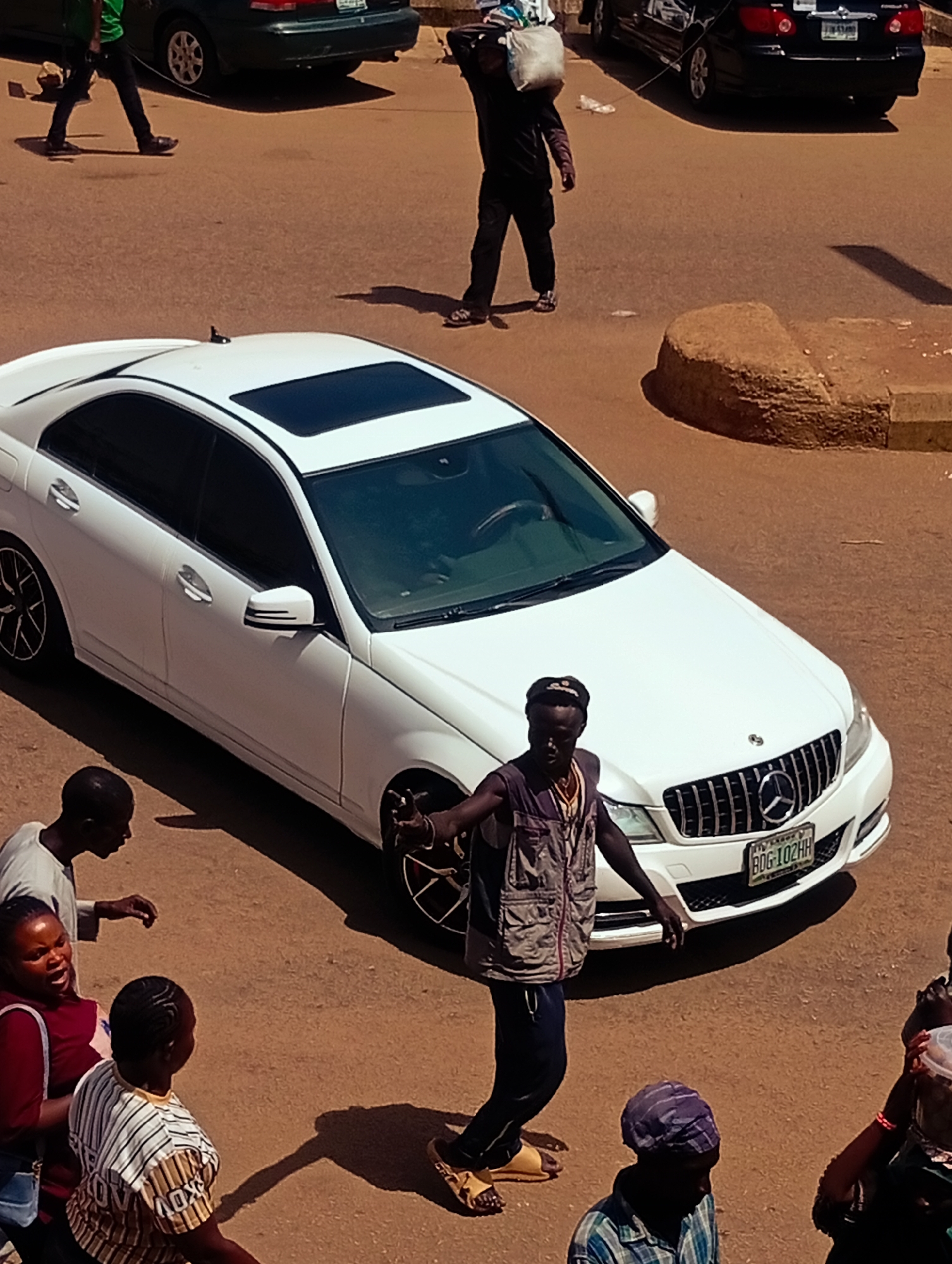 The image depicts a street scene in Bukuru, Nigeria, with several people and vehicles. In the foreground, a white Mercedes-Benz sedan is parked, facing right, with its sunroof open. A dark-skinned man in a vest and dark pants, wearing yellow sandals, stands near the front of the car, gesturing with his right hand. To the left of the car, a dark-skinned woman in a red top and blue jeans is visible, her mouth open as if speaking. Next to her, a child wearing a striped t-shirt looks away. Behind them, a dark-skinned man in a white t-shirt is seen from the side.
In the background, a dark-colored sedan is parked further down the street. A dark-skinned man in a black jacket and pants is walking away from the viewer, carrying a white bag over his left shoulder. Another dark-colored vehicle is parked on the left side of the image, and a person in a green shirt is walking in front of it. The ground is a reddish-brown, suggesting a dusty or lateritic surface. A concrete curb with a rough, textured surface is visible to the right of the white car. The overall lighting suggests it is daytime.