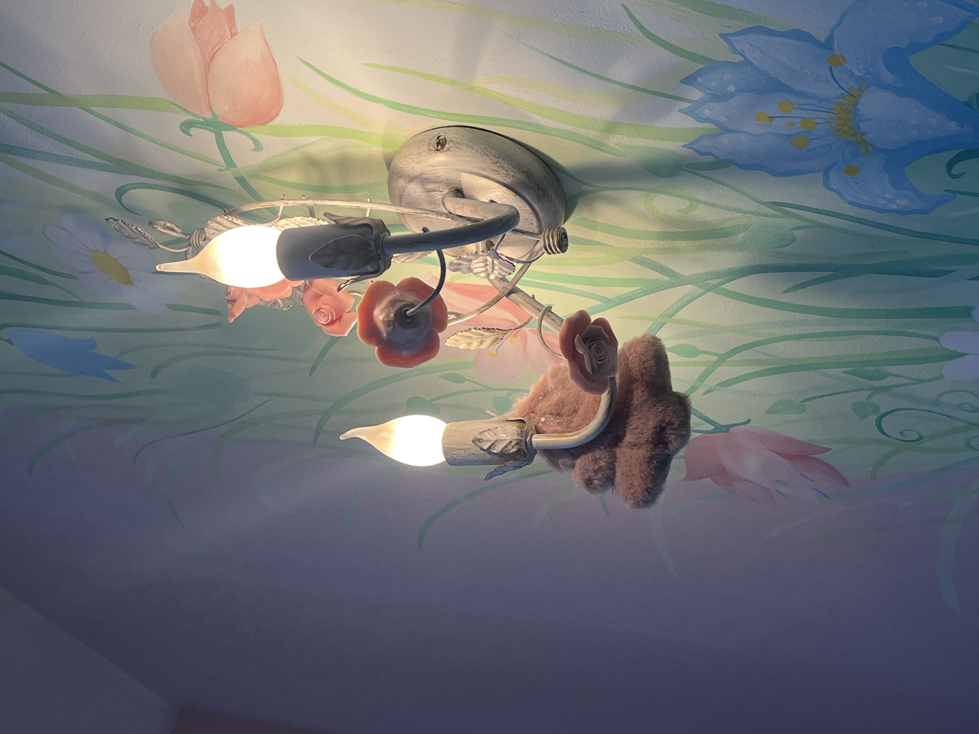 A ceiling-mounted light fixture is the central focus of the image. The fixture is adorned with flowers and fuzzy brown attachments, suggesting a whimsical or childish design. Two bulbs are illuminated, casting a warm glow. The ceiling itself is painted with a mural of various flowers and greenery in soft pastel colors, creating a whimsical, indoor, domestic environment. There are no people in the frame, so there are no actions, or interactions to describe. The city and location are unknown.