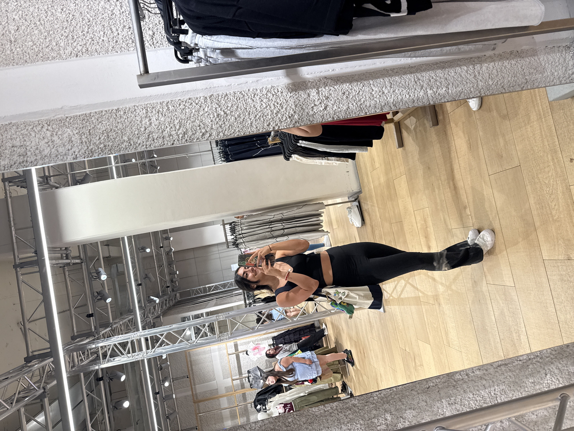 The image shows a reflection in a mirror inside a clothing store. A young woman in a black crop top and black leggings is taking a selfie. In the reflection, other shoppers can be seen browsing clothing racks. The store has wooden floors and clothing racks filled with apparel. Overhead are exposed beams with lighting fixtures. There is no information in the image to conclude that the location is Puebla, Mexico.