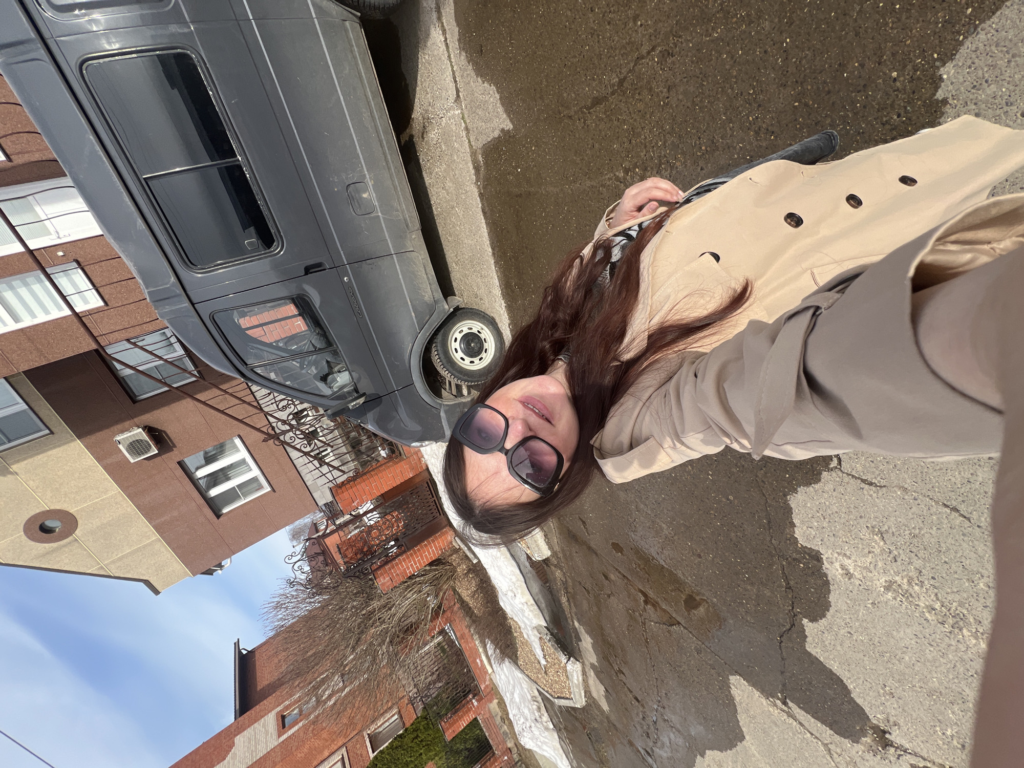 A woman wearing sunglasses and a beige trench coat stands on a wet sidewalk in Noviy, Russia. A gray van is parked nearby, adjacent to buildings of mixed brick and stucco construction, with architectural details including wrought iron railings. The sky is partly clear.
