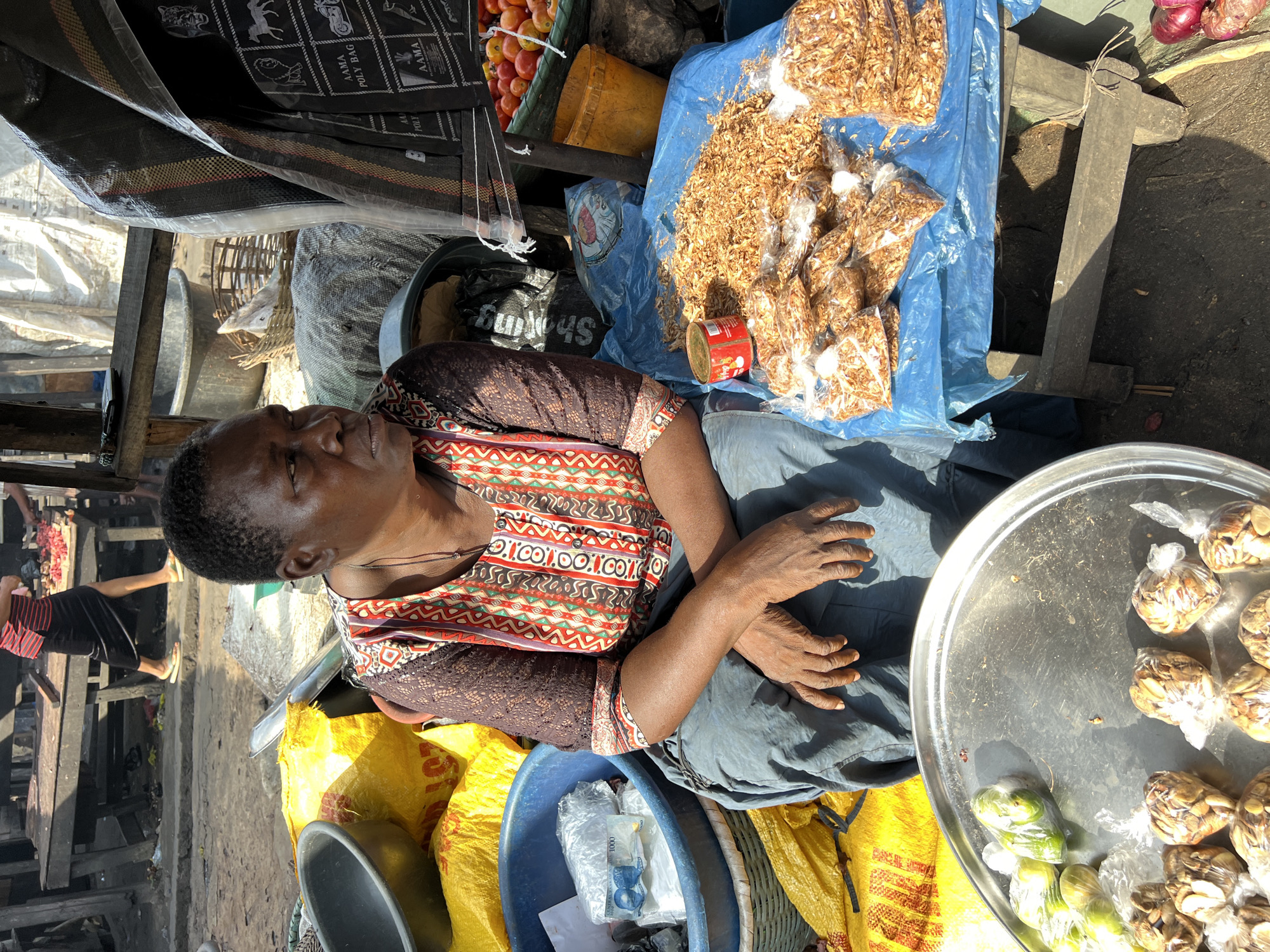 In Nkpor, Nigeria, a woman sits at a market stall, her hands resting on her lap. She wears a colorful, patterned blouse with a brown lace overlay on the sleeves and a dark blue skirt. In front of her, on a blue tarp covering a wooden stool, are several clear plastic bags filled with dried shrimp and a small red can. To her left, a large blue basin contains items wrapped in plastic, including what appear to be bank notes. A silver tray to the far left holds several small, individually wrapped food items. Behind the woman, yellow bags are piled high, and a variety of baskets and containers are visible. The market appears to be outdoors, with sunlight casting shadows on the ground. In the background, a person in a red and black striped shirt and black skirt walks past a wooden market table laden with raw meat.