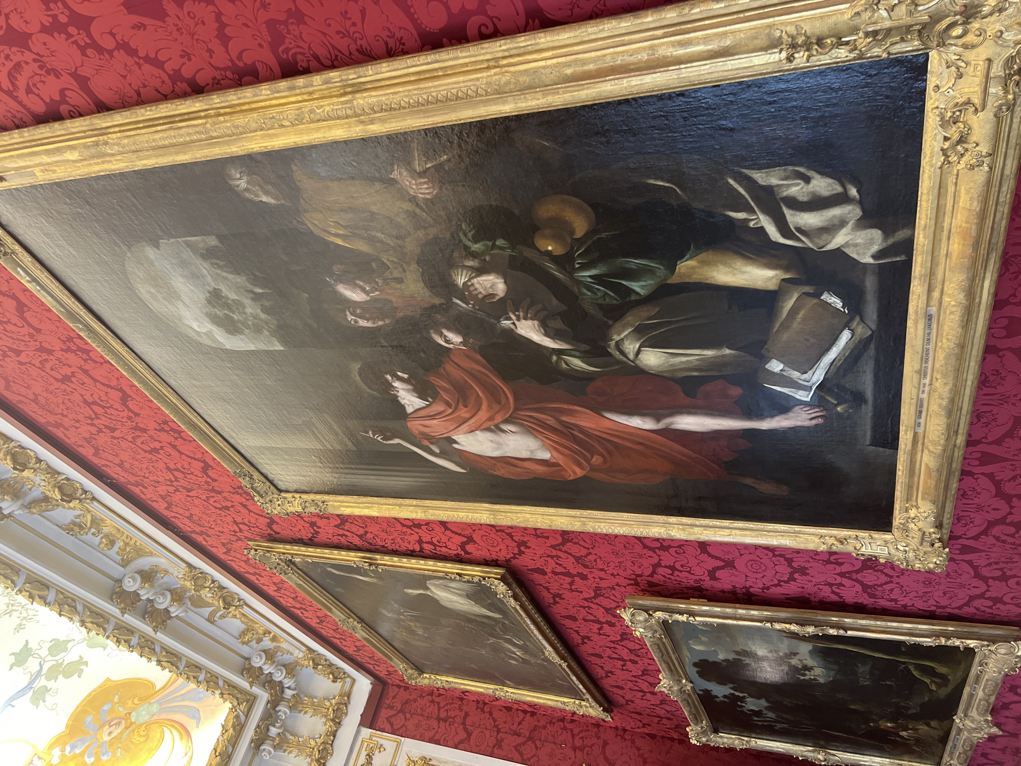 The image shows an opulent room with red patterned wallpaper and ornate gold trim on the walls and ceiling. Three framed paintings are visible on the wall. The largest painting in the foreground depicts a religious scene, likely a biblical event, with a central figure in a red cloth reaching out to a man in a green garment. Several other figures are present in the background. To the left of this large painting, a second, smaller painting shows a landscape scene with figures and animals. Further to the left and higher up, a third painting displays a bird in a dark, wooded setting. The ceiling above is decorated with intricate gold moldings and a fresco of a winged figure. The overall impression is of a richly decorated art gallery or a formal living space within a historical building. The location is the Schleissheim Palace, in Oberschleissheim, Germany, specifically in the New Palace (Neues Schloss).