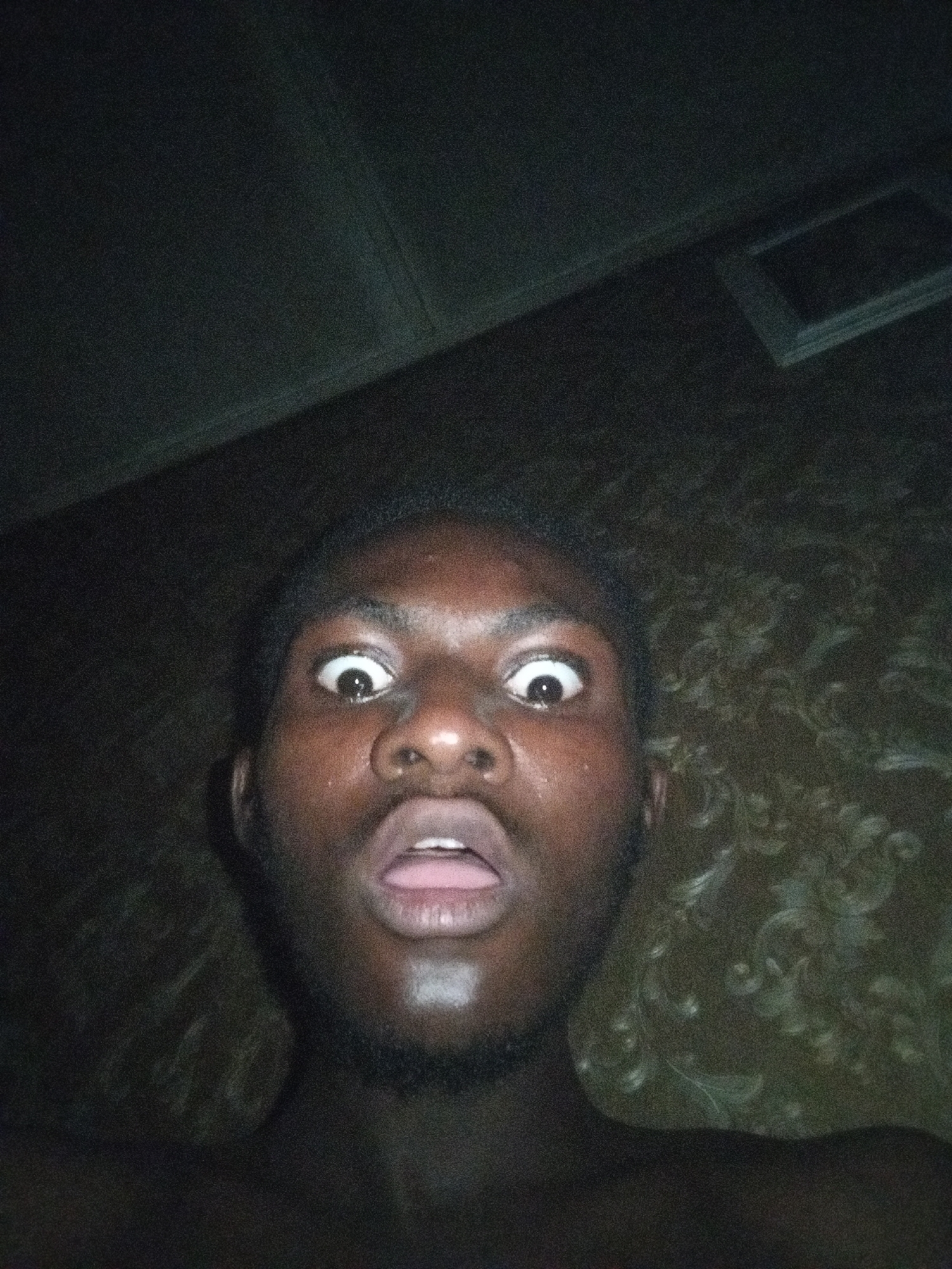 The image shows a dark-skinned person with wide eyes and an open mouth, positioned close to the camera. The lighting is dim, revealing a patterned surface behind them, possibly wallpaper. The person appears to be shirtless. The location is stated as Unknown, Unknown.